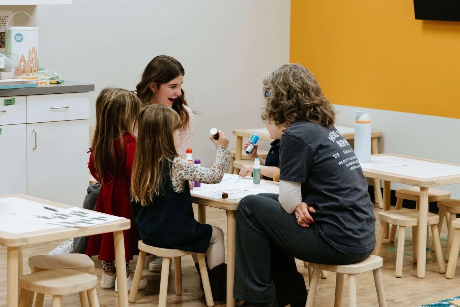 A woman and four young girls sitting at a table in a classroom or activity room, engaging in arts and crafts with bottles of glue and craft materials. The woman is smiling and interacting with the children.