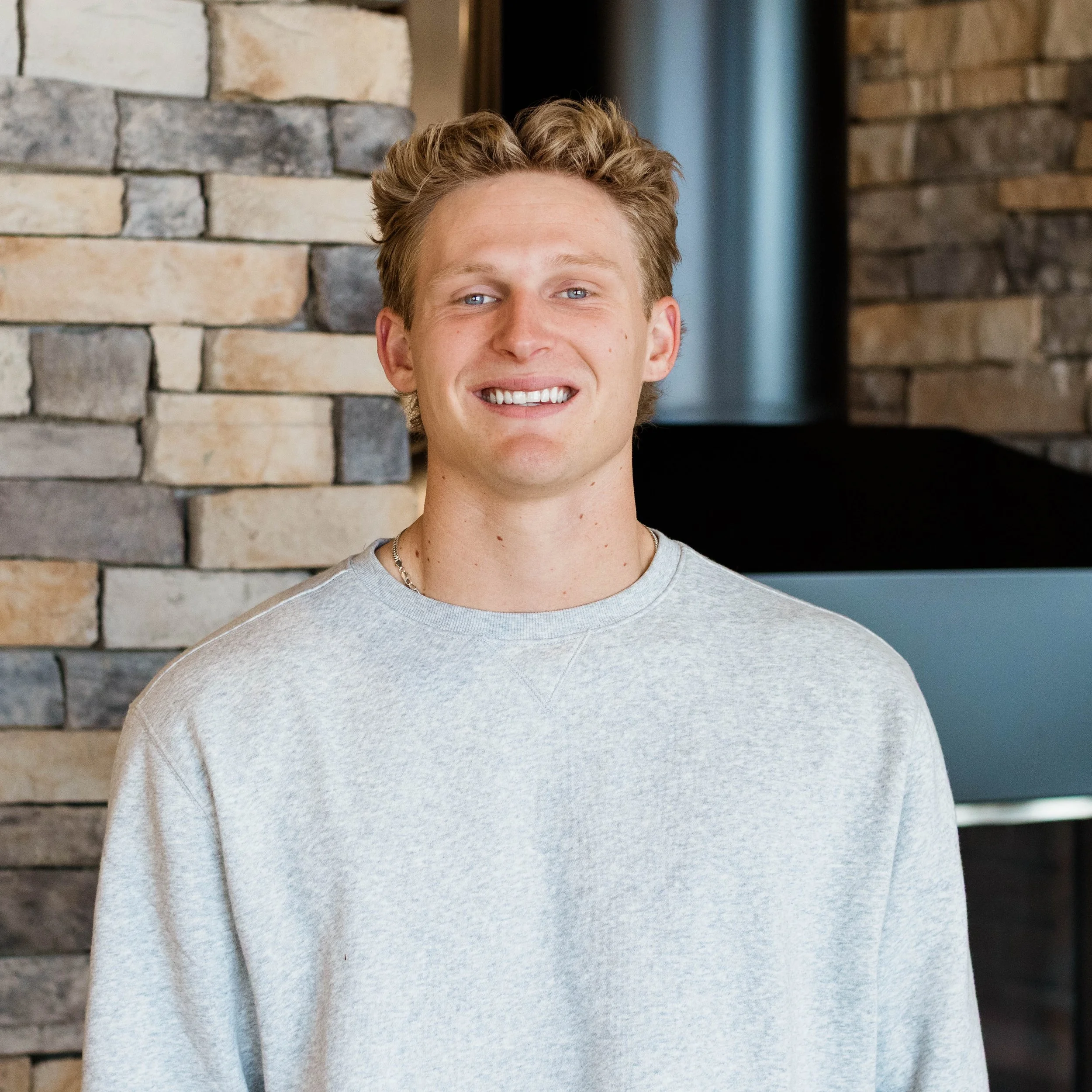 A young man with curly red hair and blue eyes, smiling, wearing a light gray T-shirt, standing indoors near a brick wall.