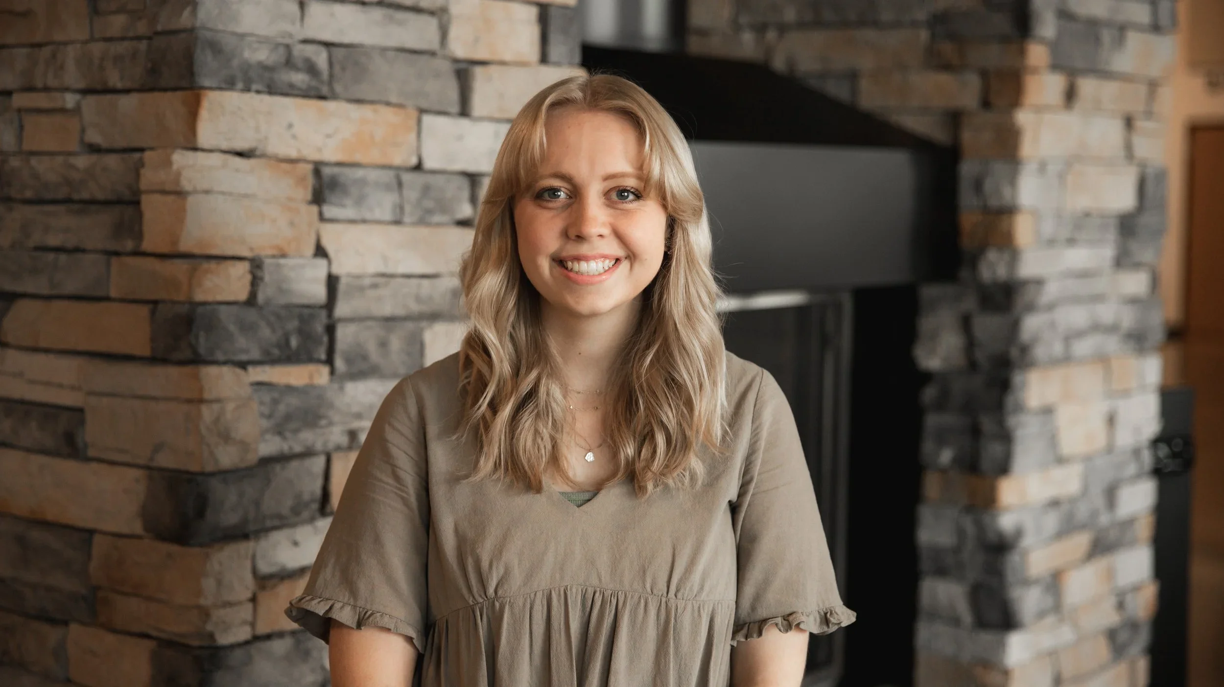 A young woman with long, wavy blonde hair smiling indoors, standing in front of a stone fireplace.
