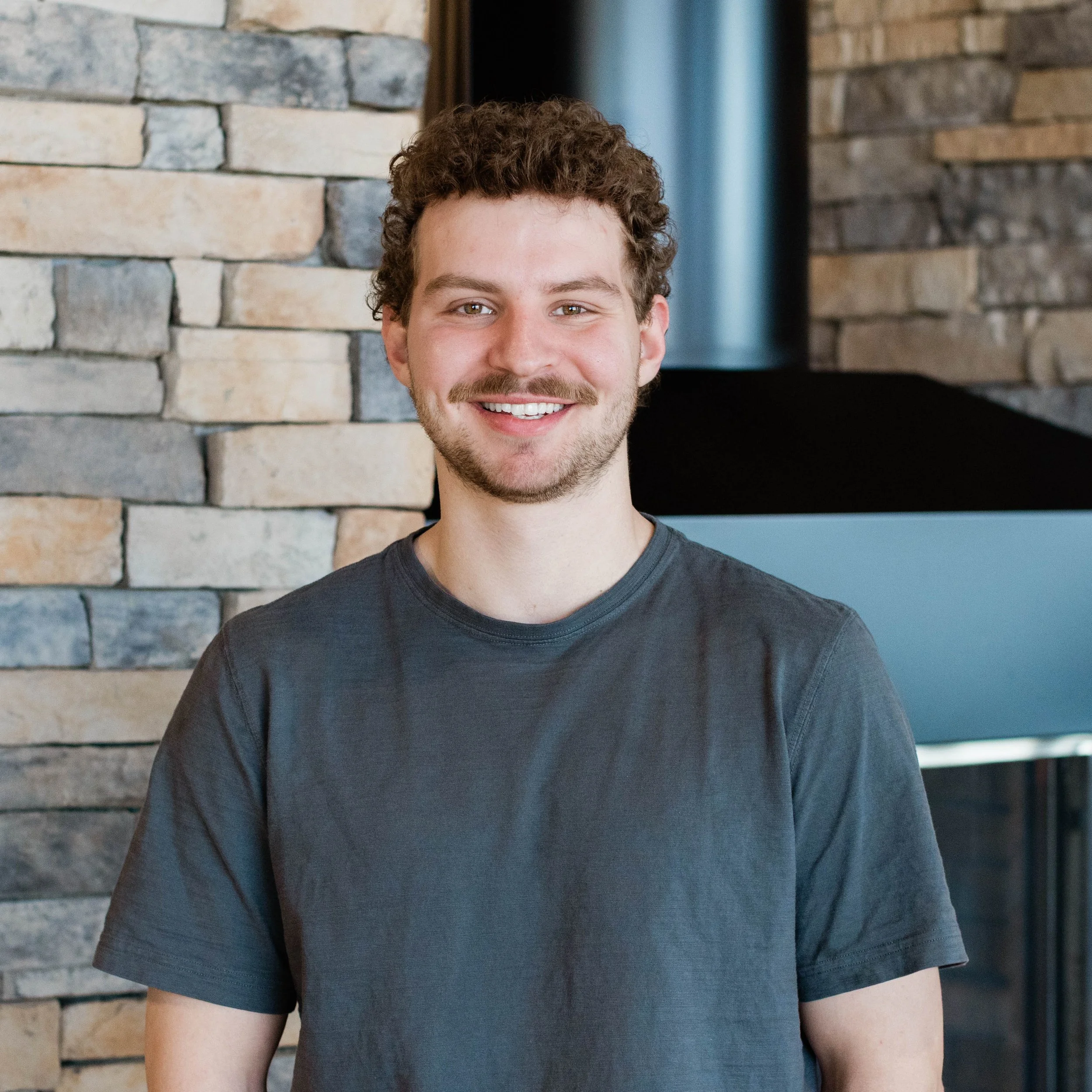 A smiling young man with curly brown hair and a mustache, wearing a black Carhartt T-shirt, standing indoors in front of a stone fireplace.