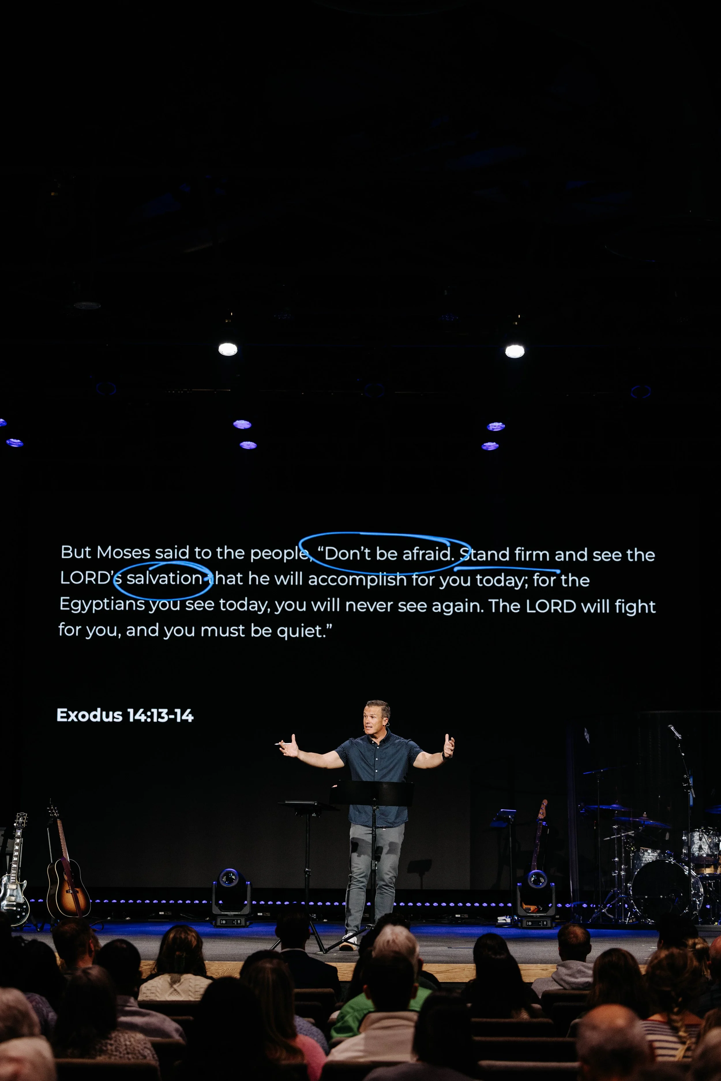 Crowd attending a Christmas concert with Christmas trees and decorations, stage with performers, and large screen.