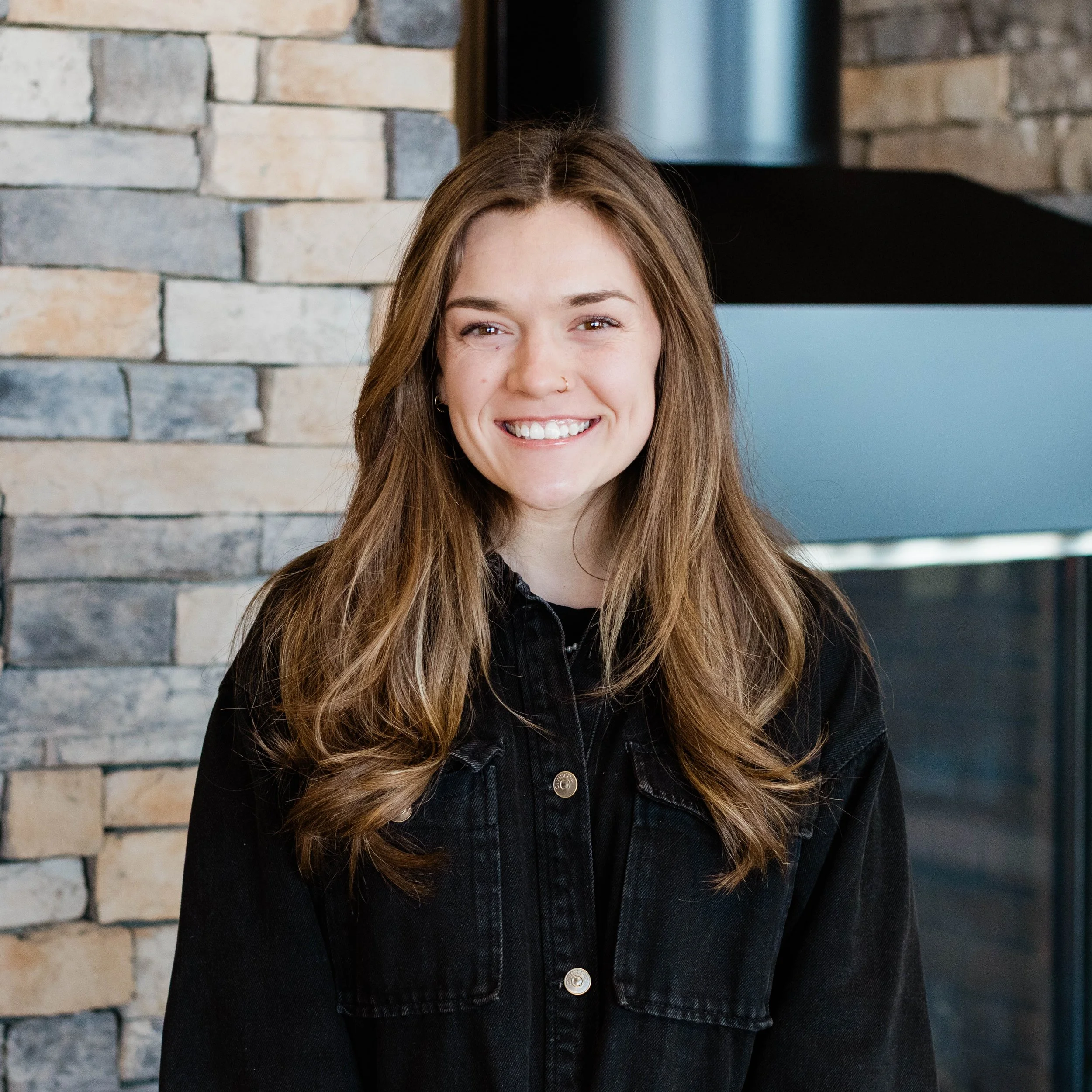 A young woman with long wavy brown hair smiling, wearing a black jacket over a white shirt, standing in front of a stone wall with a fireplace.