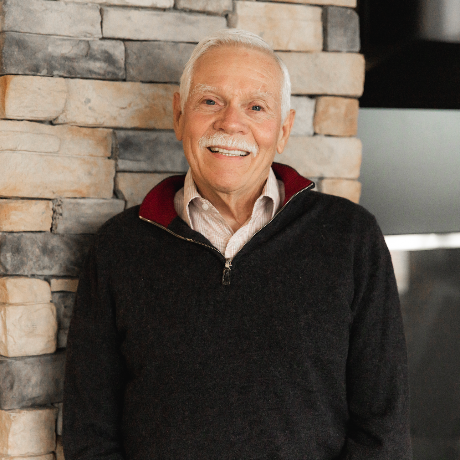 An older man with white hair and a mustache smiling, dressed in a dark sweater over a light-colored shirt, standing in front of a stone wall.