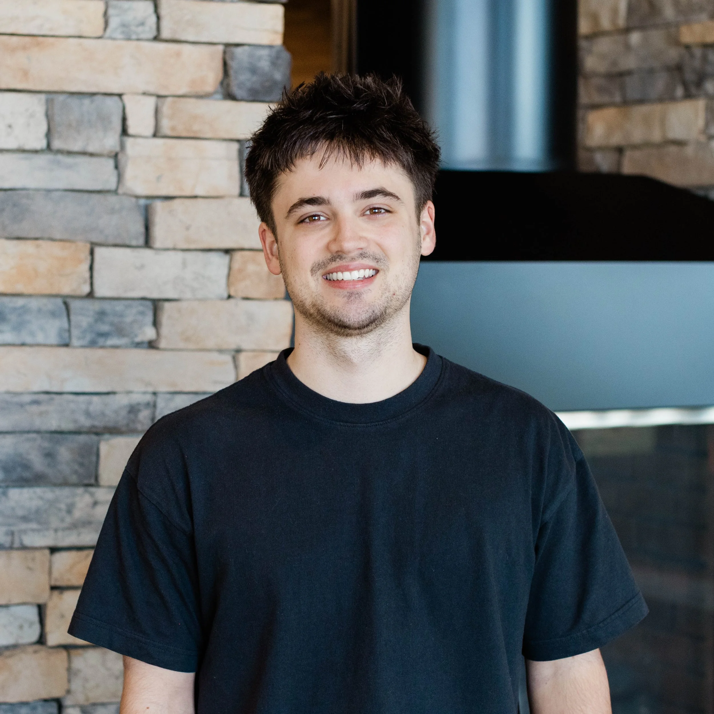 A young man with short dark hair, wearing a plain white t-shirt, smiling in front of a stone wall interior.