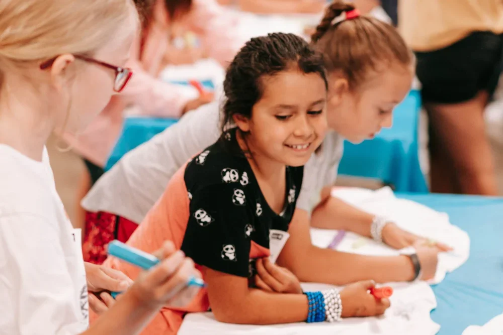 A group of children leaning over a table, engaging in an activity, with a young girl smiling and wearing a black shirt with skull patterns.