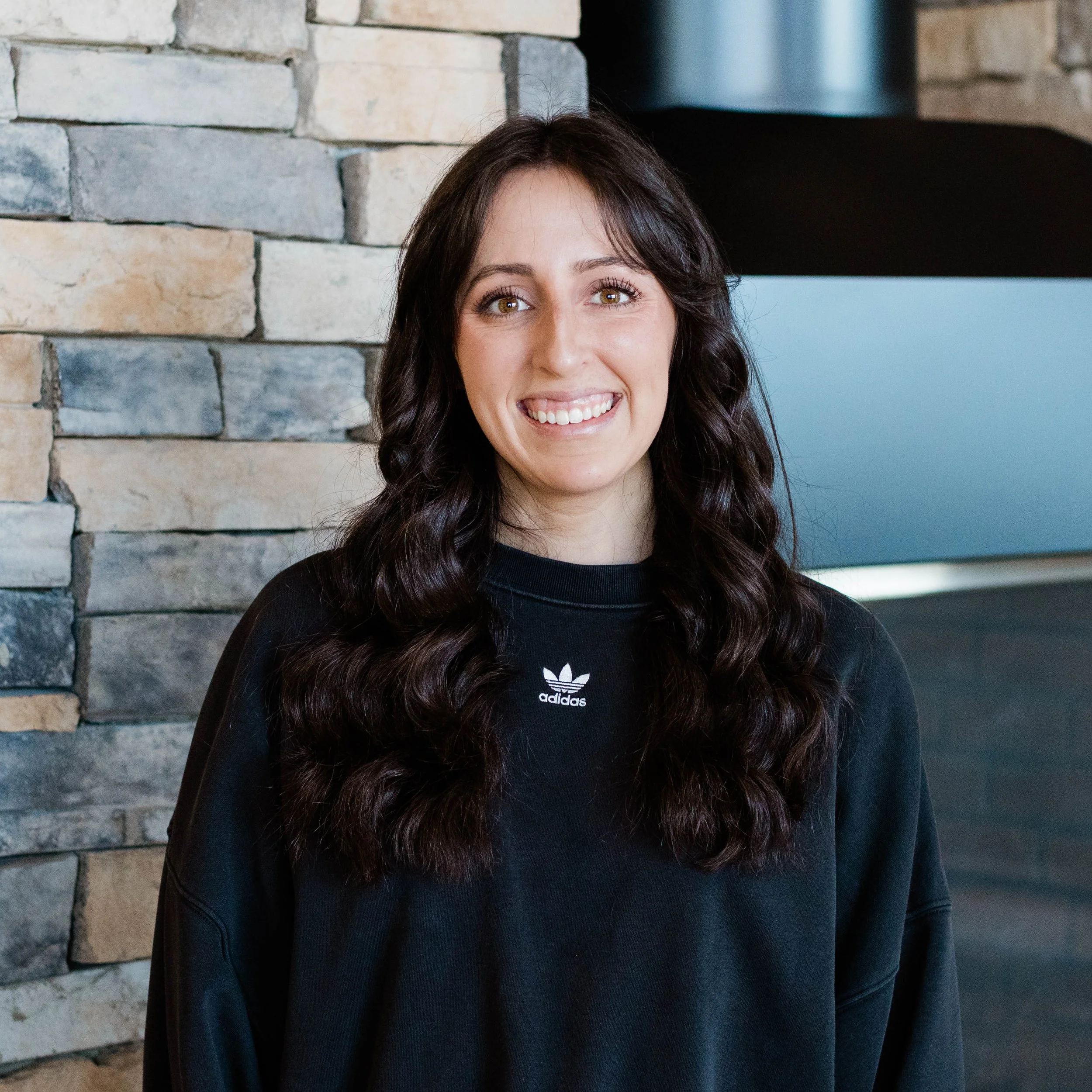 A woman with long, curly dark hair smiling in front of a stone fireplace.