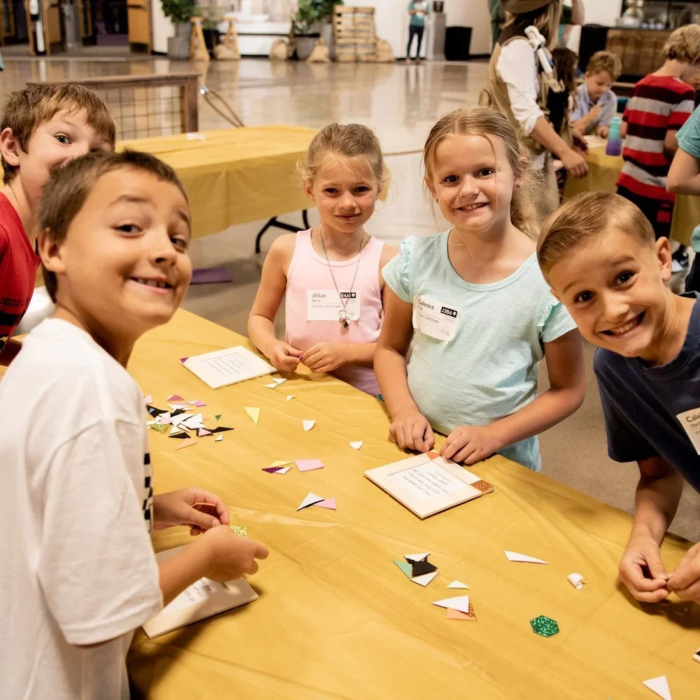 Group of five children smiling and working with paper crafts at a table indoors.
