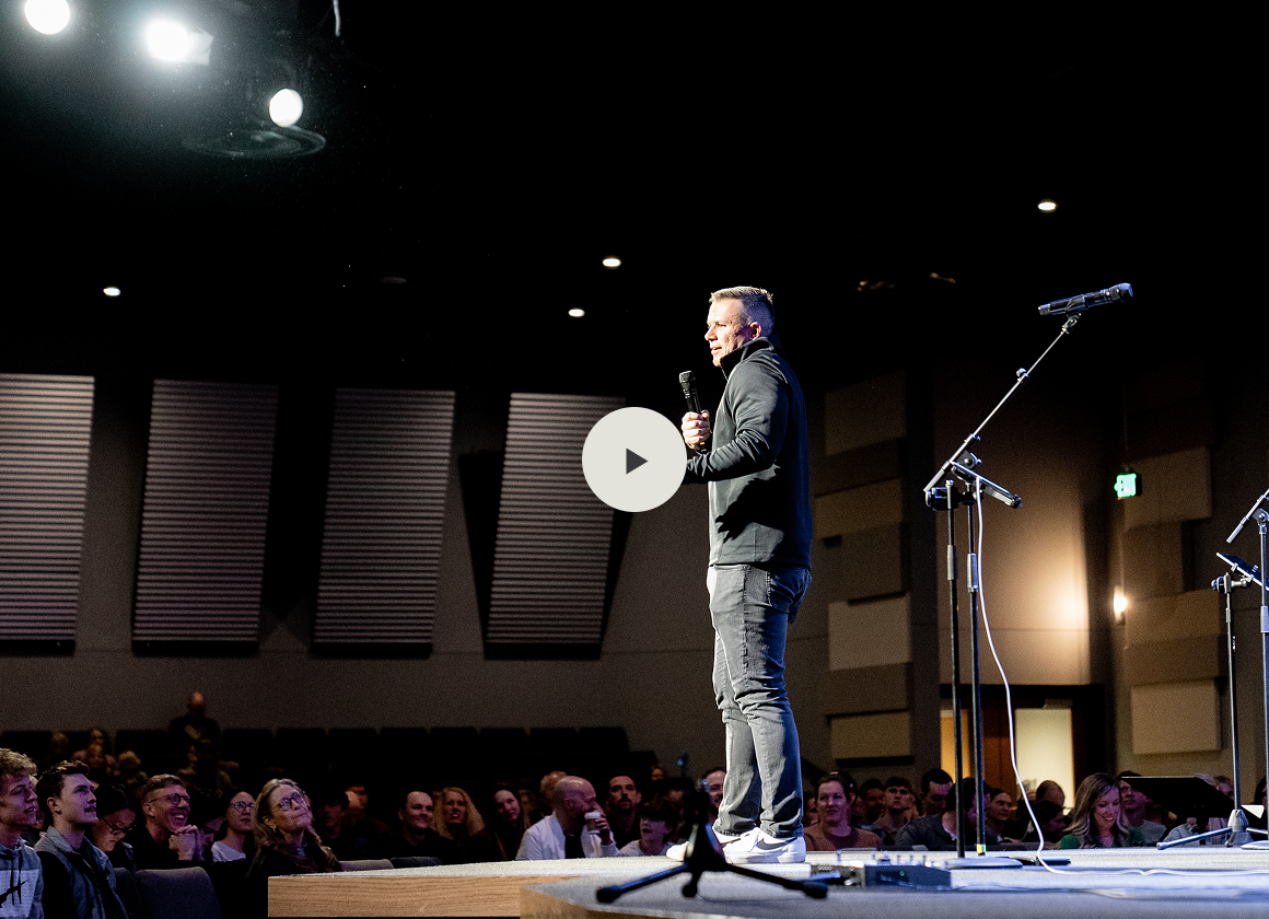 A man standing on a stage holding a microphone in front of a seated audience in a dimly lit auditorium.