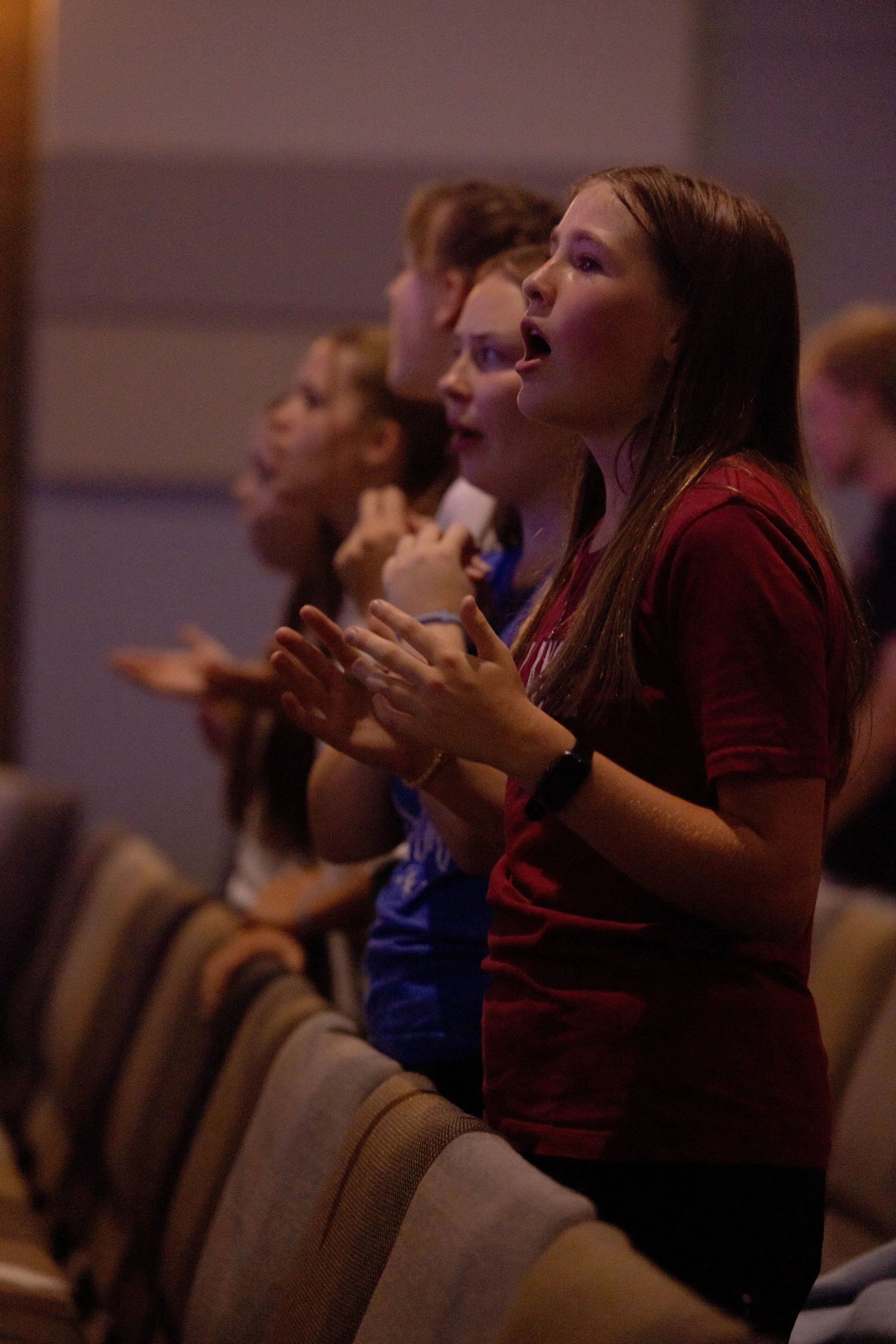 Group of young people standing and singing or speaking during an indoor event, with chairs in the foreground.