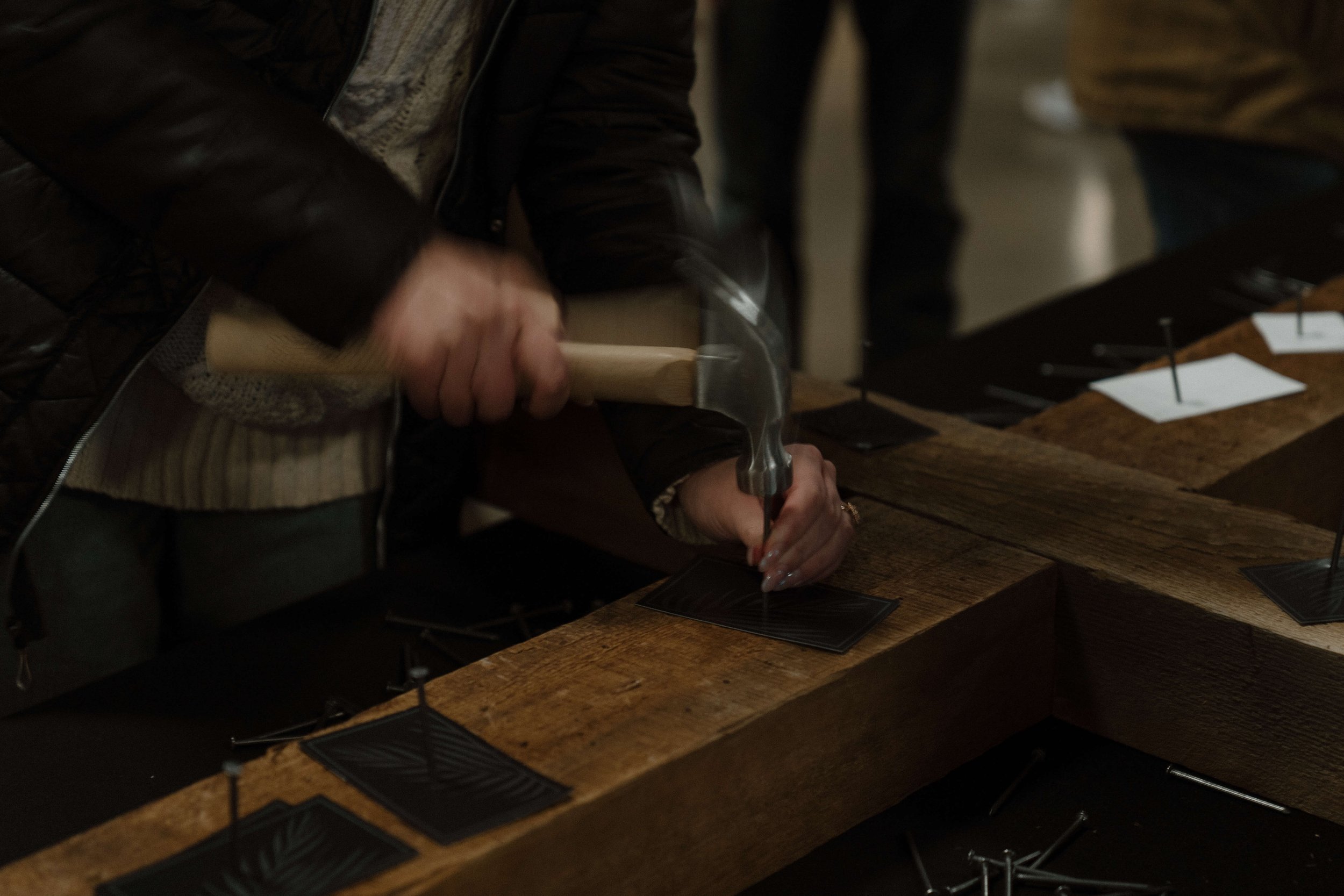 Person hammering nails into a wooden table in an indoor setting, with other hands and nails visible on the table.