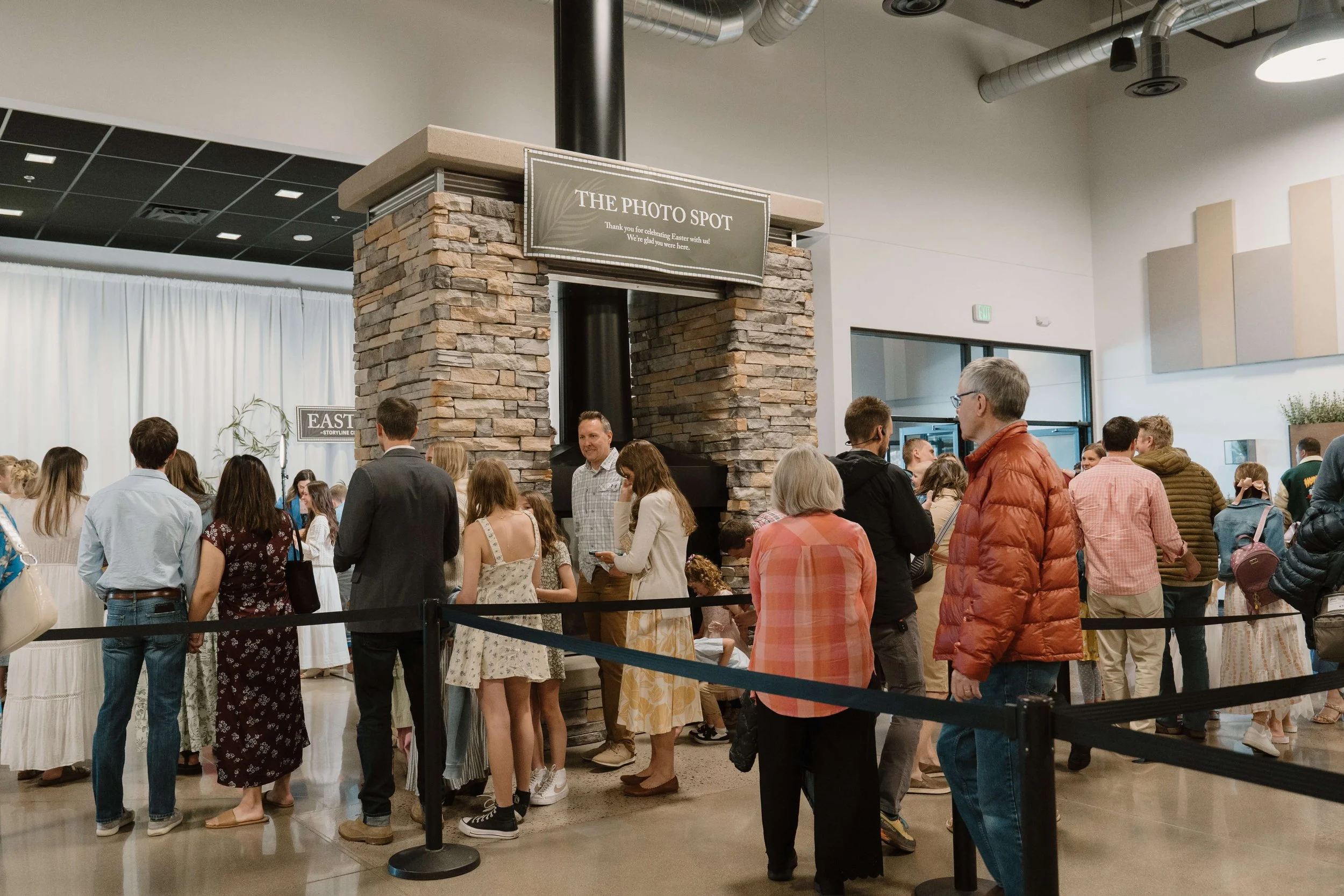 People standing in line inside a church, near a stone fireplace with a sign that reads 'The Photo Spot,' taking photos because of Easter celebration.
