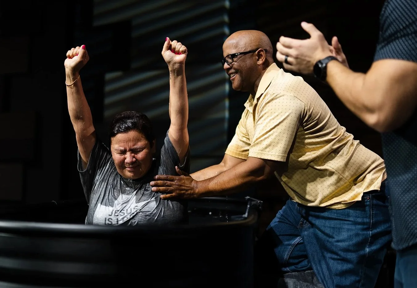 A woman is being baptized, surrounded by people, with one man assisting and another clapping.