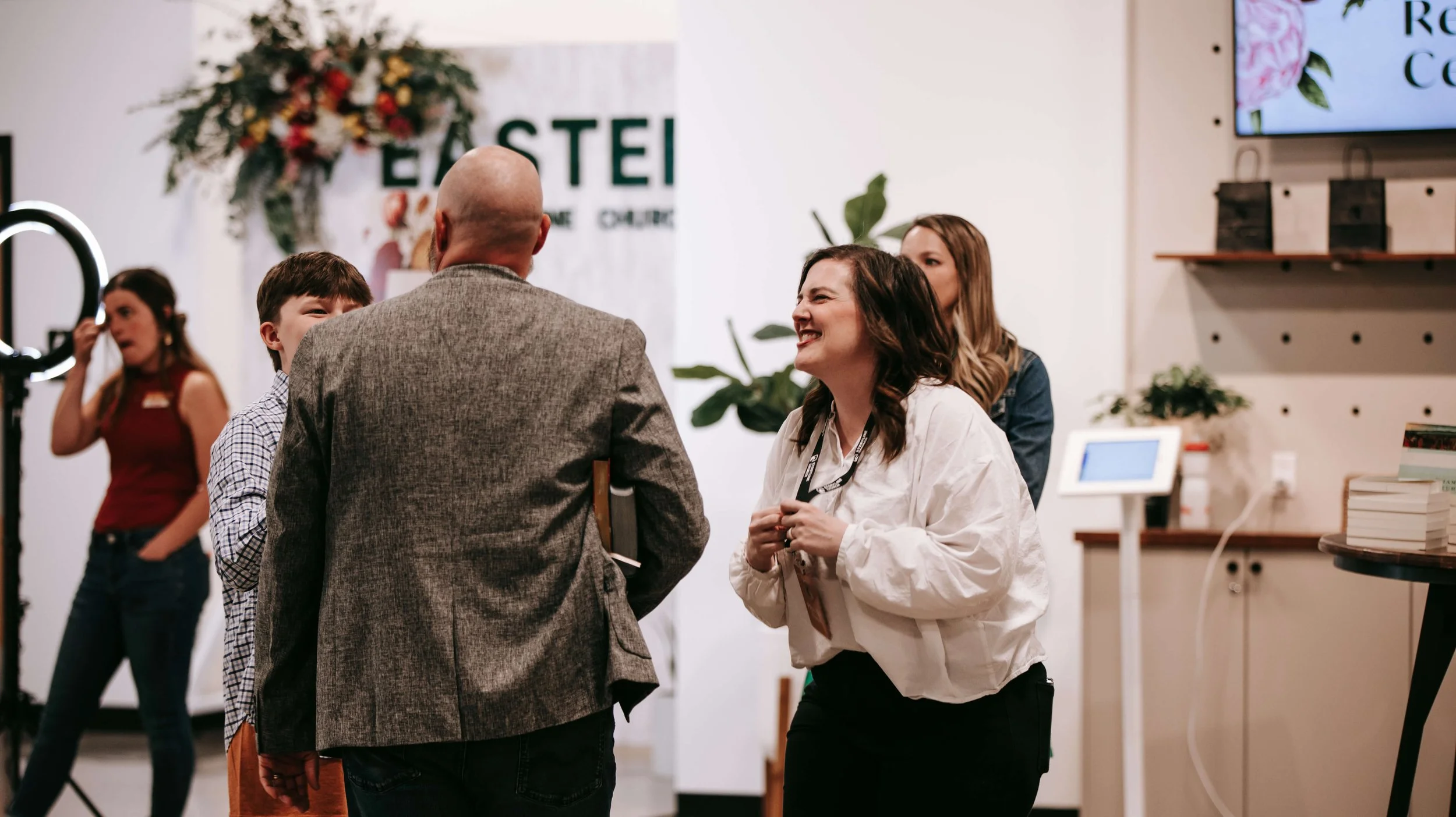 People socializing at an indoor event, with a large floral display and a sign that reads "EASTEL" in the background.