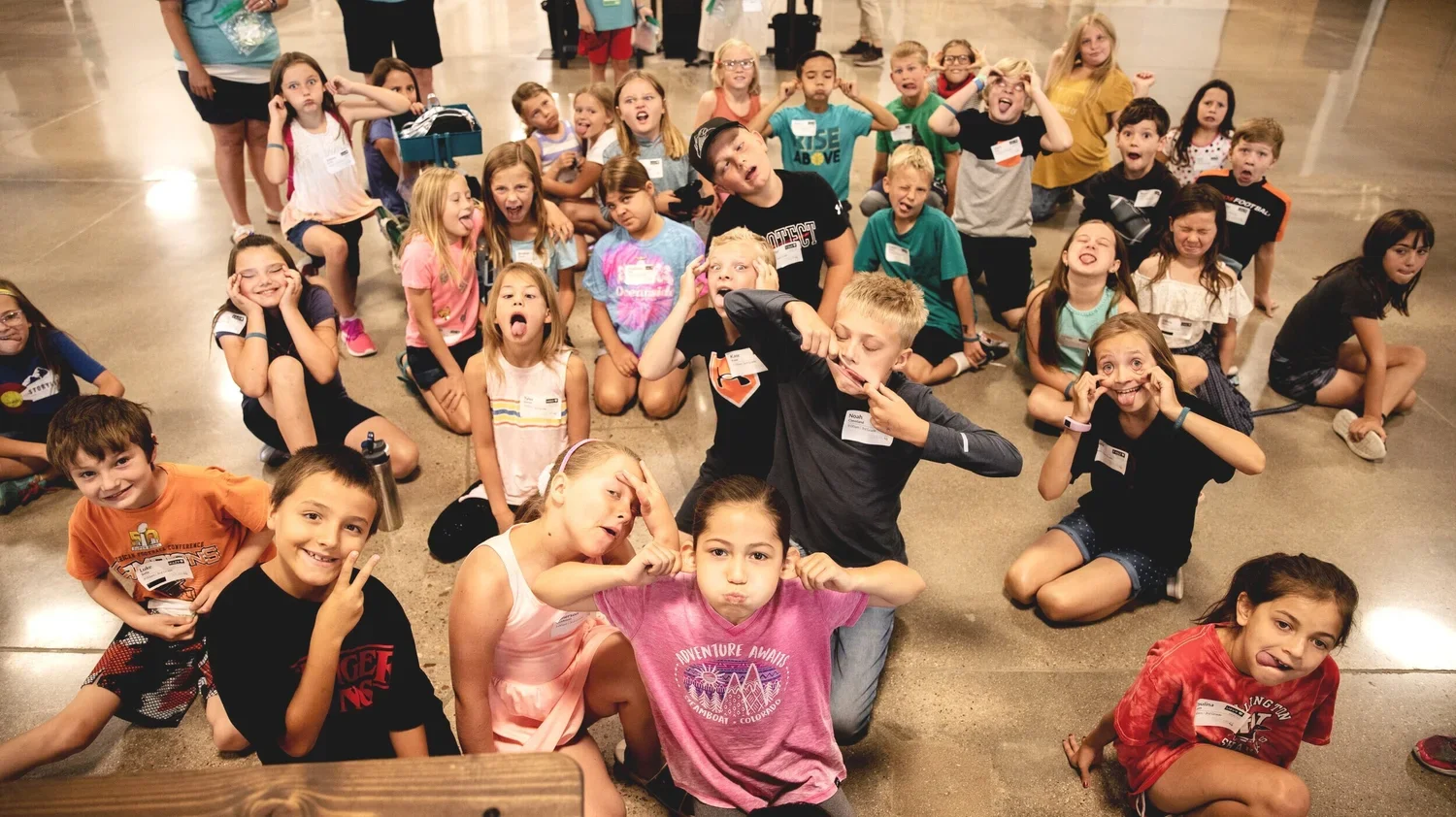 Group of children sitting and kneeling on the floor, making silly faces and gestures, inside a large indoor space.