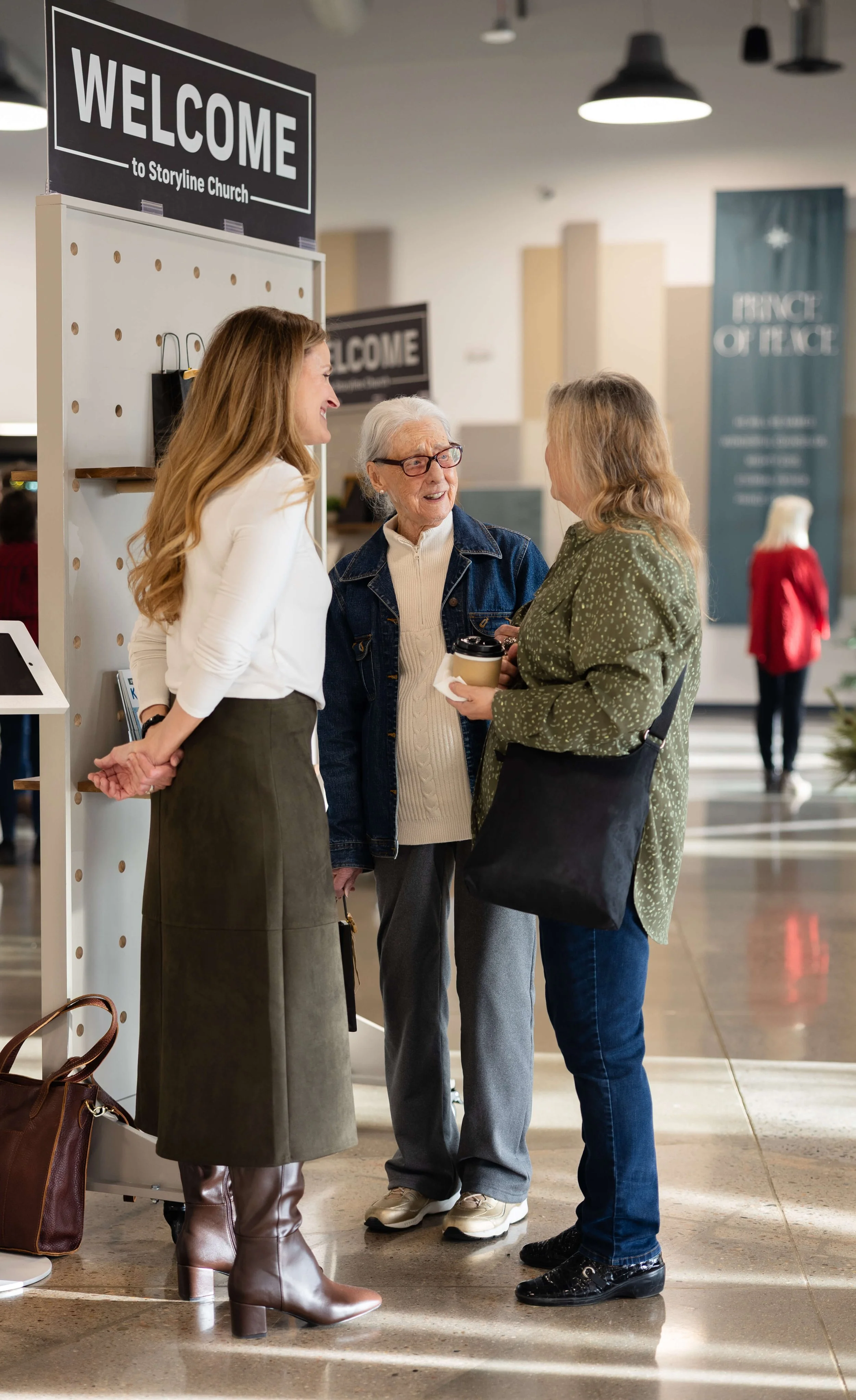 Three women having a conversation near a sign that says 'Welcome to Storyline Church' inside a modern church lobby.