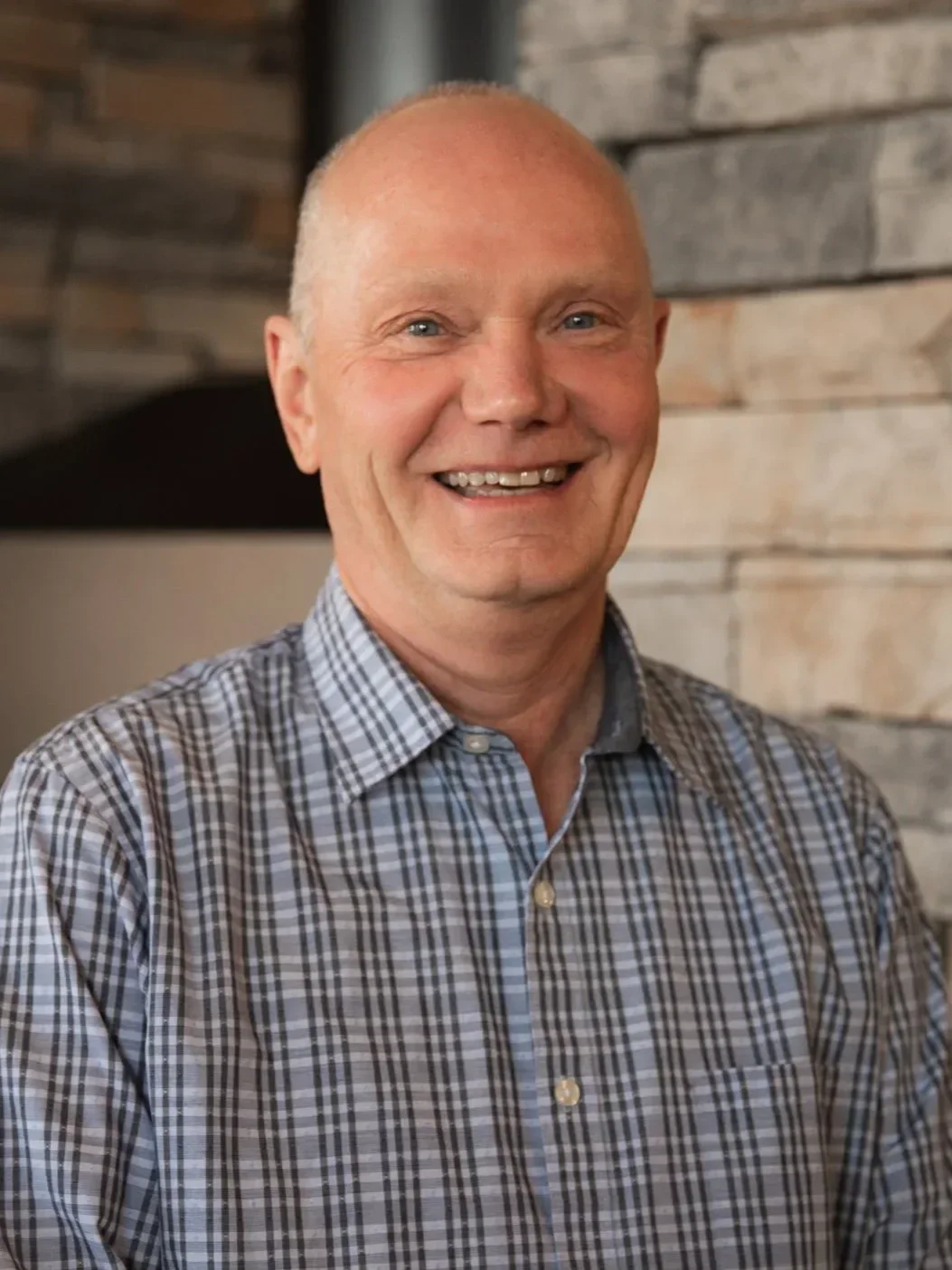 A smiling middle-aged man with a bald head wearing a checkered shirt standing against a wooden wall background.