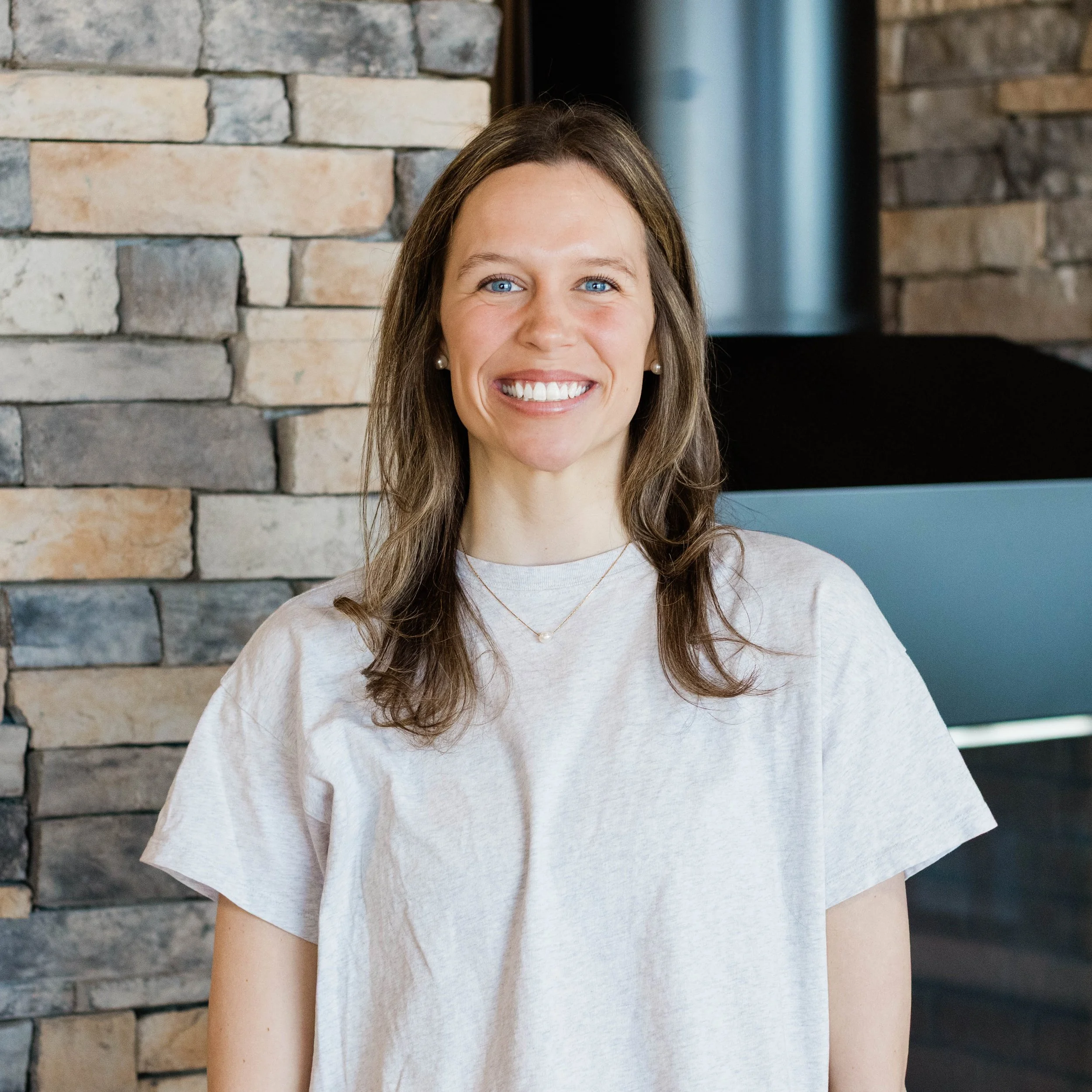 A woman with shoulder-length brown hair, smiling, wearing a light blue T-shirt, standing indoors in front of a stone wall with a fireplace.