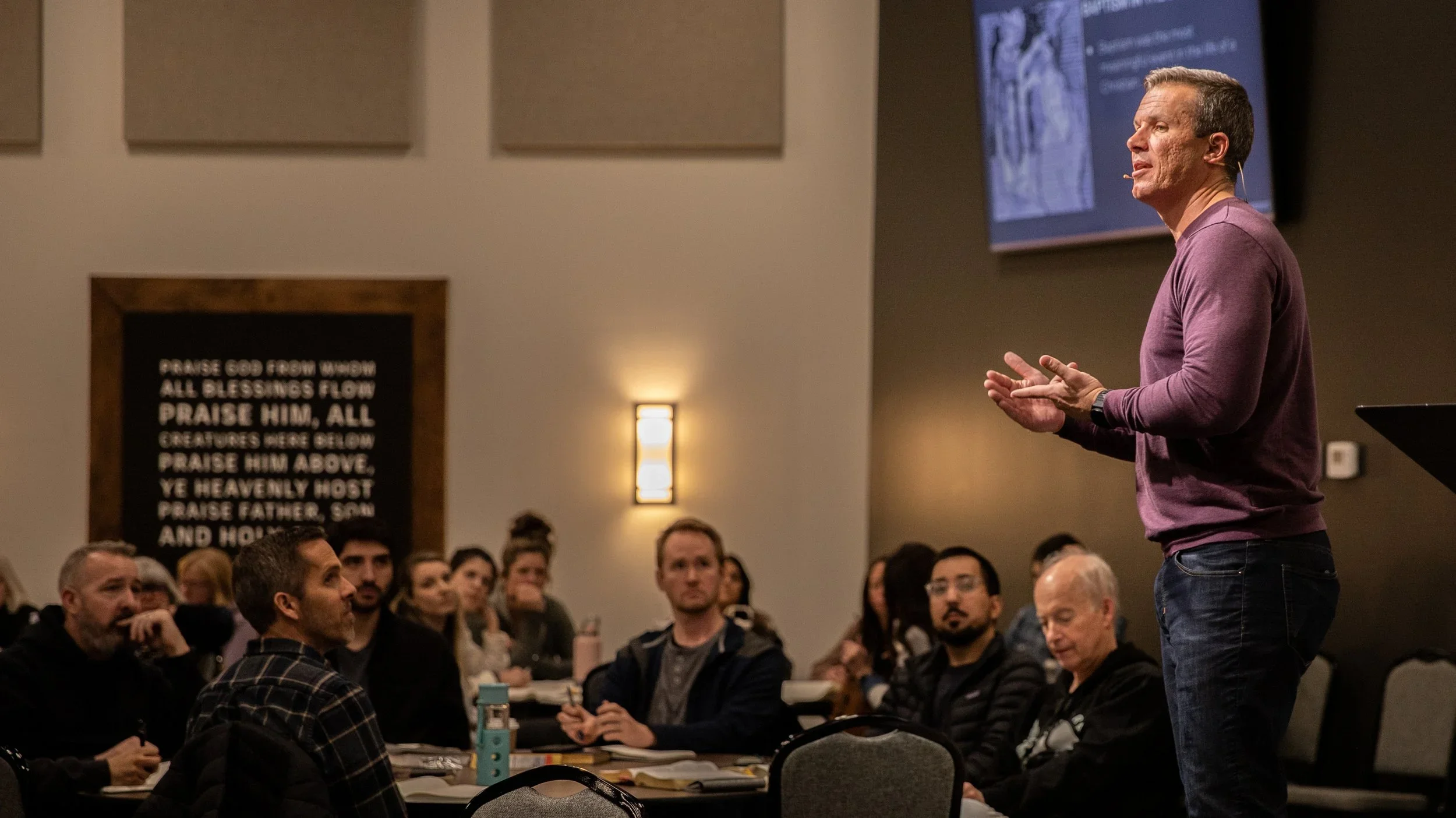 A man in a purple long-sleeve shirt speaking to an audience in a conference room, with a large screen behind him and an illuminated wall sconce.