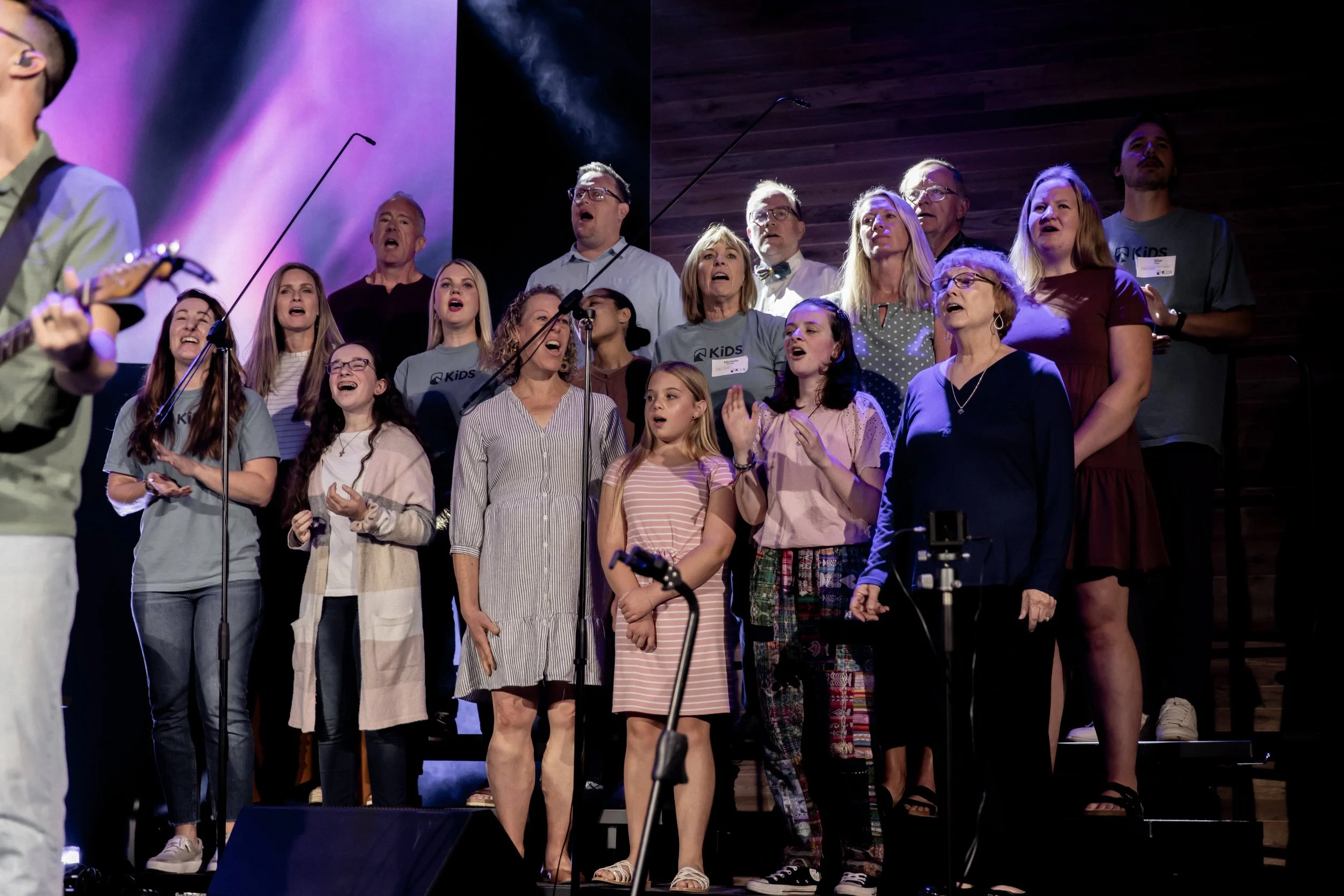 A diverse group of people, including children and adults, singing on a stage with microphones in front of a purple and black background.