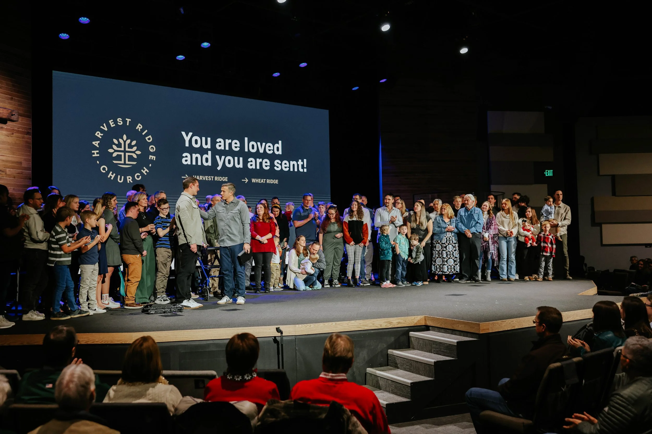 Group of people on stage at Harvest Ridge Church, including children and adults, with a large screen behind displaying the church's logo and the message 'You are loved and you are sent!'