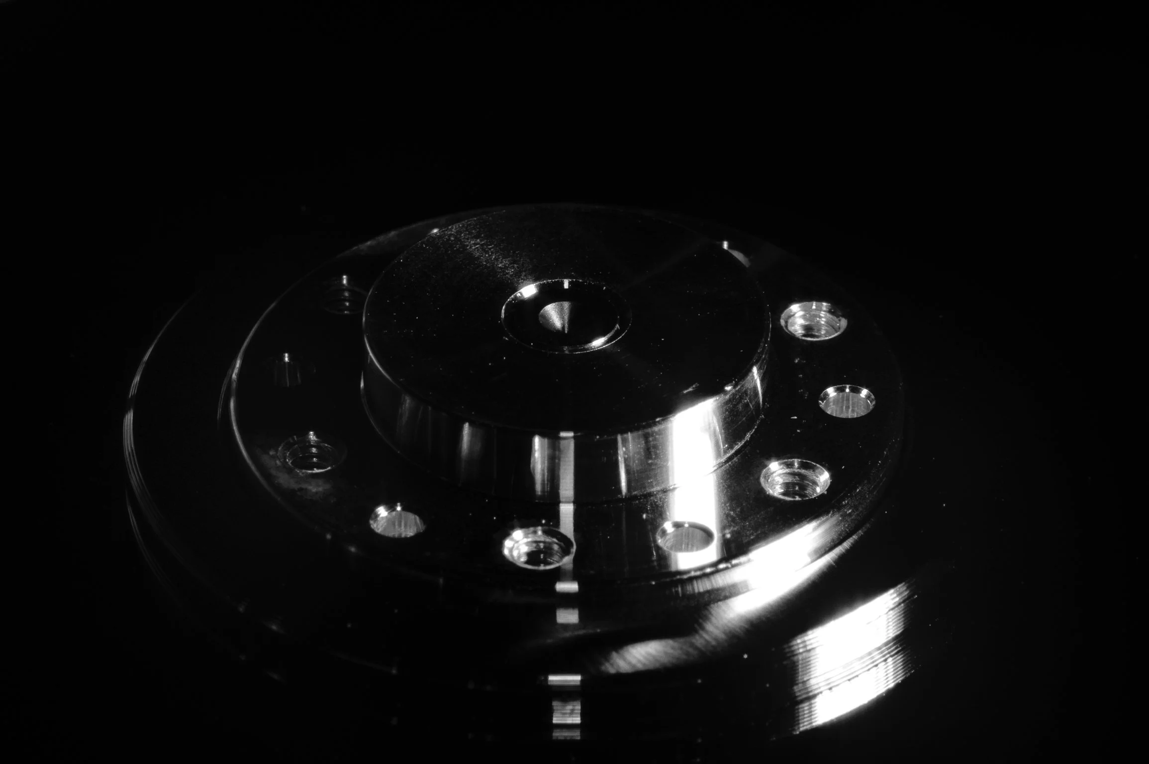 Een rond, metalen onderdeel met meerdere gaten en een centrale opening, afgebeeld op een zwarte achtergrond.