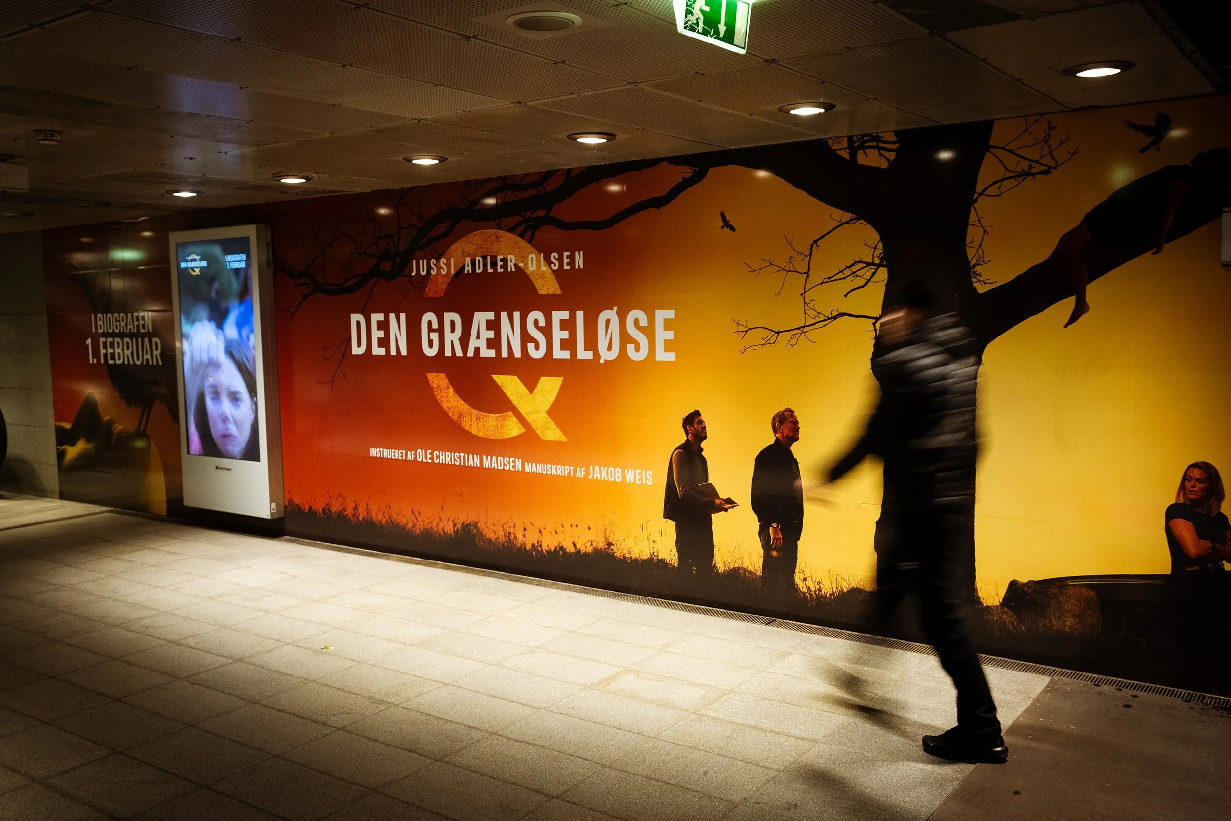 Billboard for the book "Den Grænseløse" by Jussi Adler-Olsen, with a sunset background, silhouettes of two men and a girl, and a person walking past.