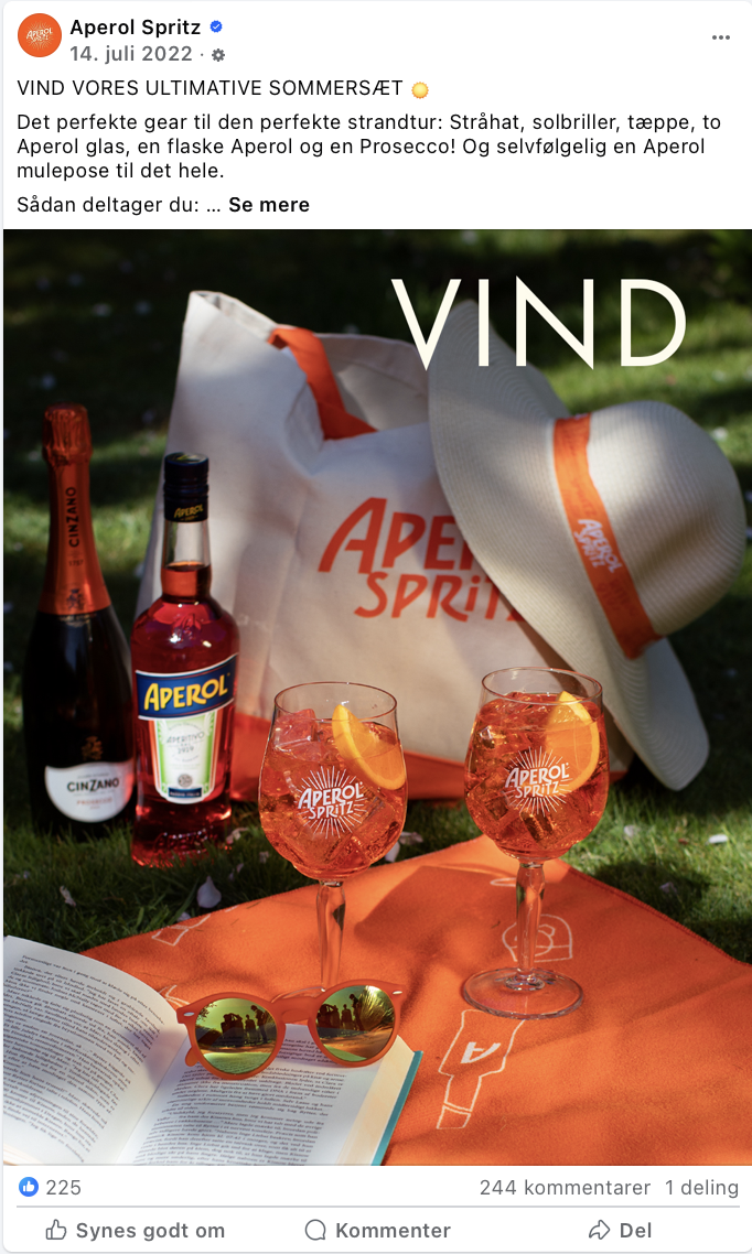 To glas med Aperol Spritz med appelsinskiver, en Aperol-flaske, en prosecco-flaske, en stor hvid strawhat og et par orange-skimede solbriller på en orange dug, med en åben bog.