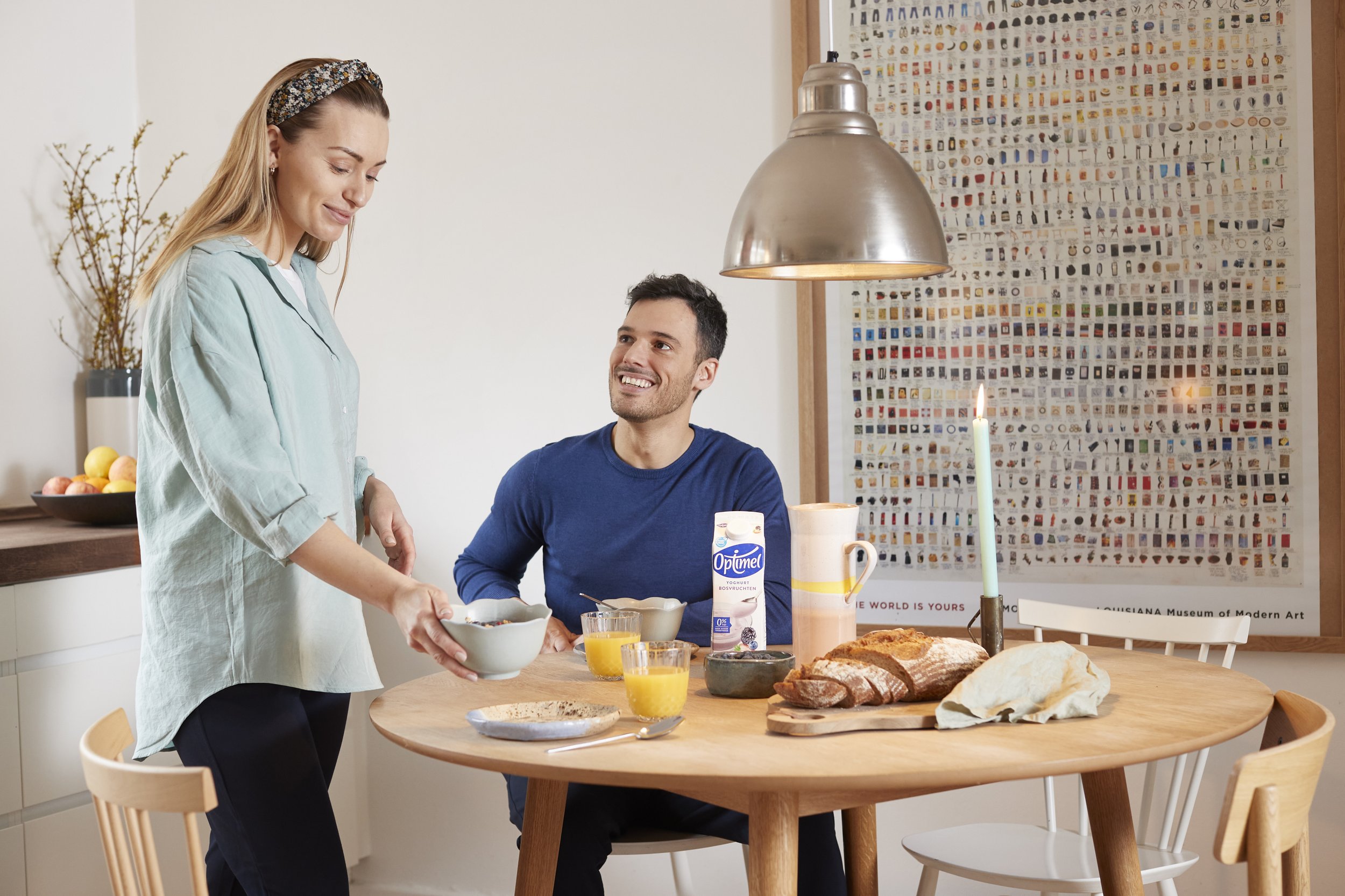 En mand og en kvinde nyder morgenmad ved et rundt træbord. Kvinden hælder noget i en skål, mens manden smiler og ser på hende. Der er brød, mælk, juice, en kop kaffe og et lys på bordet.