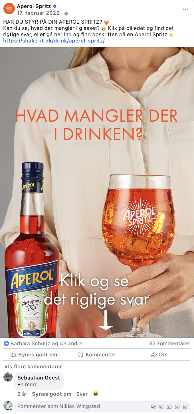 Person holder et glas Aperol Spritz med is og citrusskiver, ved siden af en flaske Aperol. Der står på billedet: 'HVAD MANGLER DER I DRINKEN?'