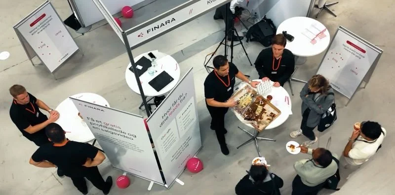 Personer står omkring bord med kage og informationstavler på en messe eller udstilling.