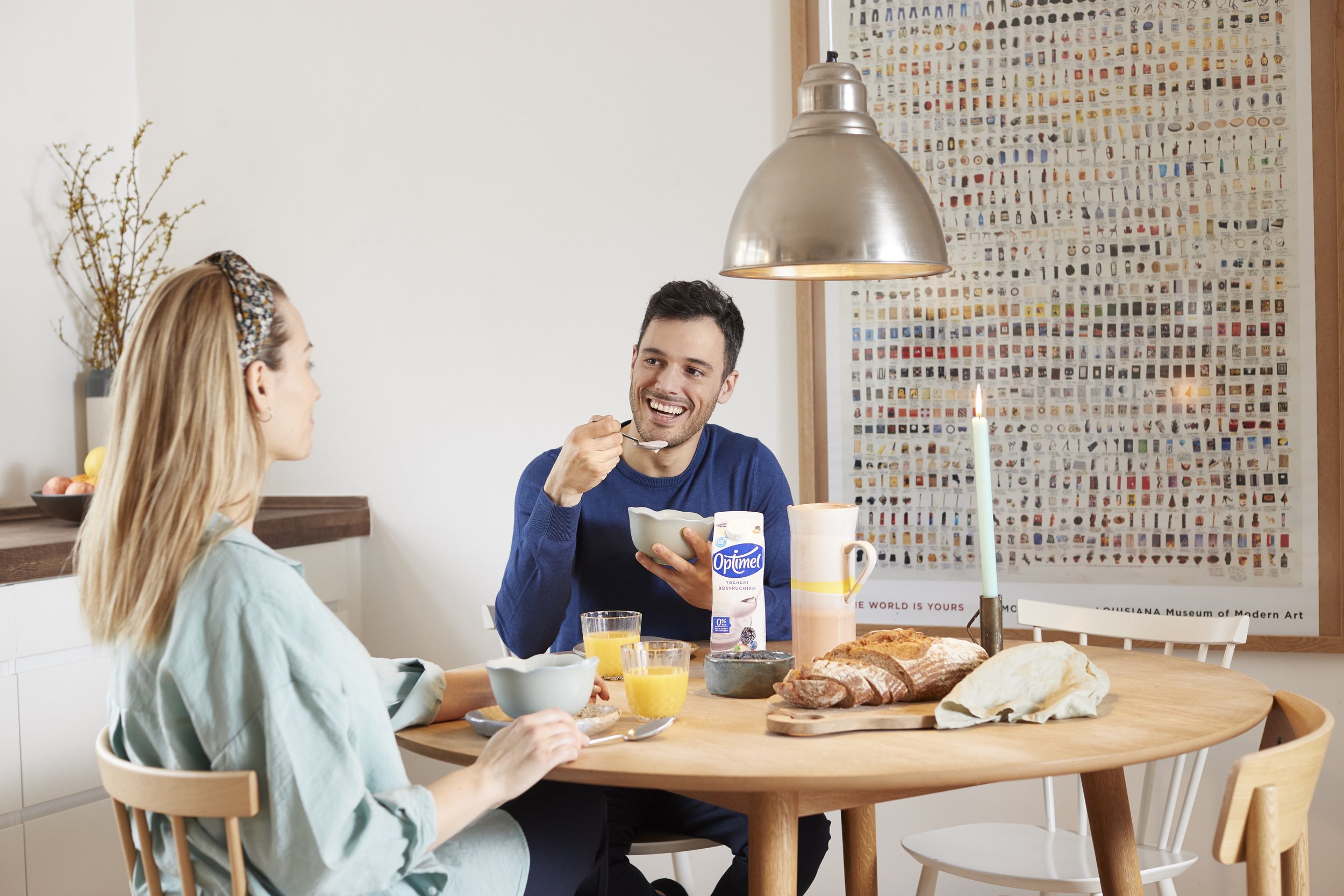To personer sidder ved et rundt spisebord og spiser morgenmad. Der er brød, et glas juice, mælk og en stearinlys på bordet. Den ene person smiler mens han spiser, den anden holder en skål