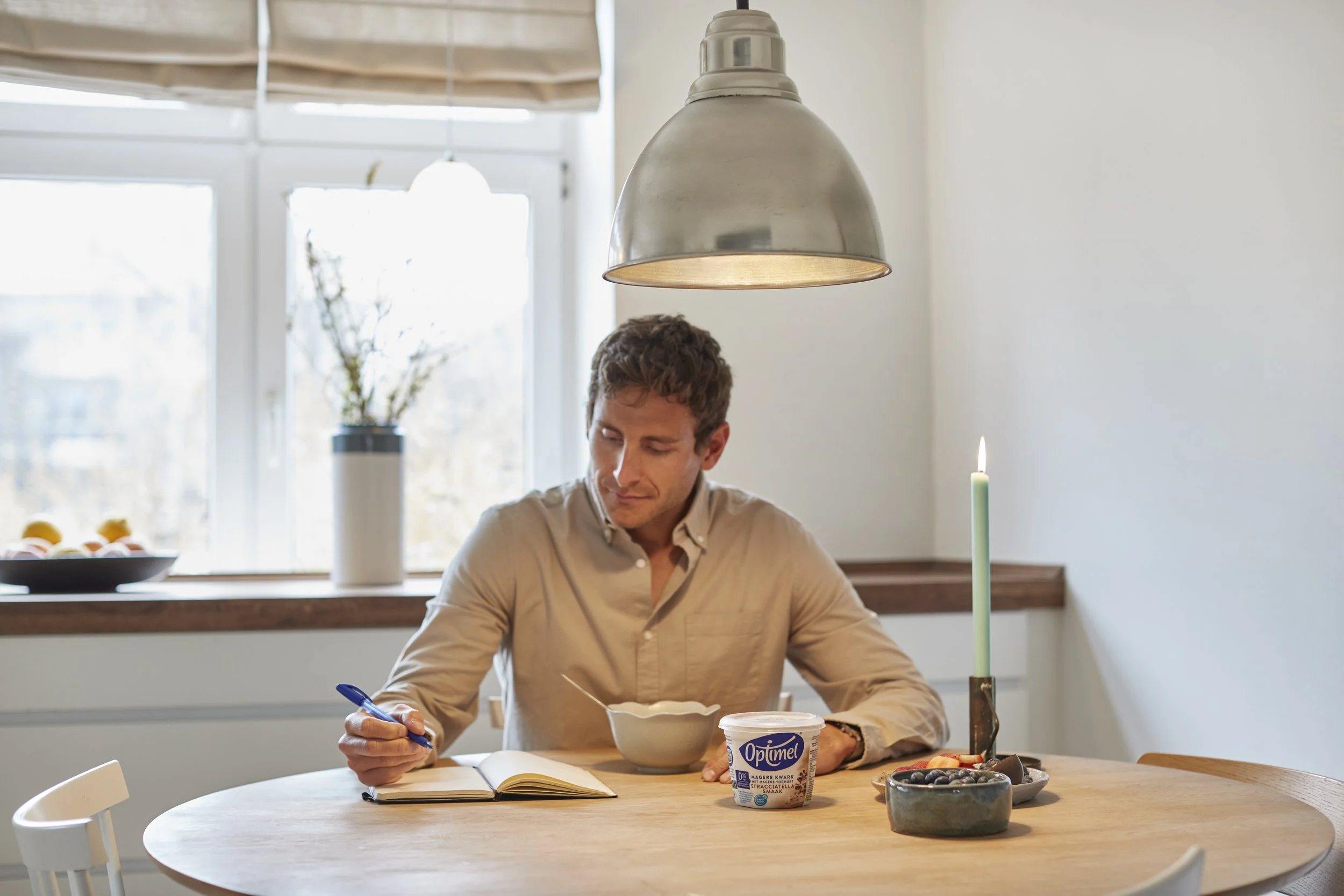 Mand sidder ved spisebord med opvask, spisegrej, og en åben bog, mens han skriver med en pen. Der er et lysestage med en grøn blomst og stearinlys, og en skål med blåbær.