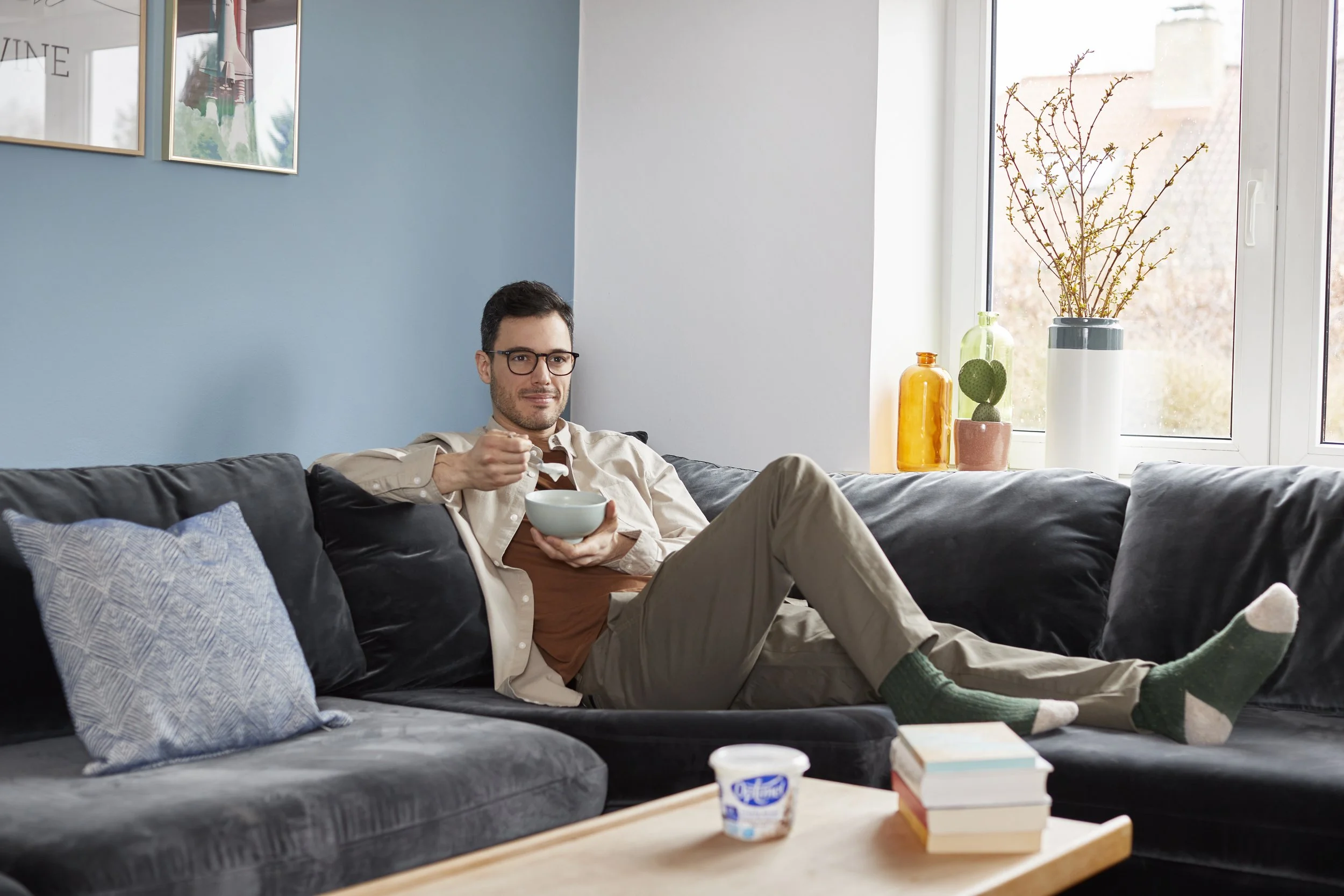 En mand sidder afslappet på en sort sofa, nyder en skål og har på sig grønne sokker. Der er et sidebord foran ham med en yoghurt og nogle bøger. Bag ham er der et vindue med blomsterdekorationer.
