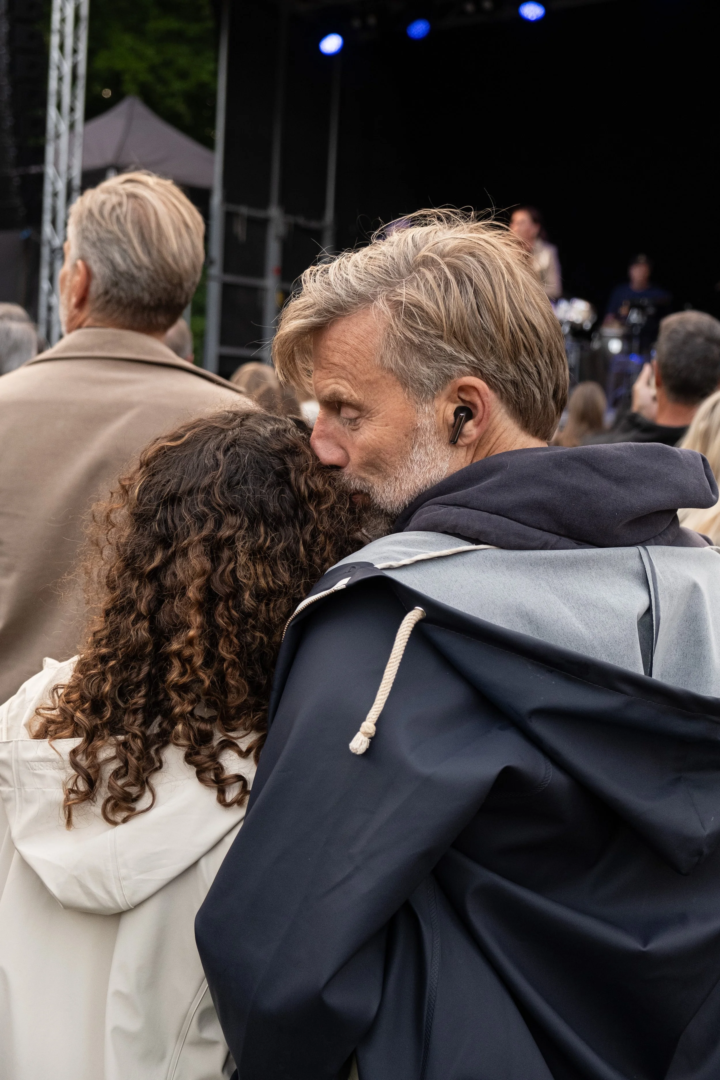 To personer står tæt sammen i en folkemængde ved en udendørs koncert. Den ene person er en ældre mand med gråt og blond hår, der har en sort trådløs øretelefon i øret, og den anden person har brunt, krøllet hår.