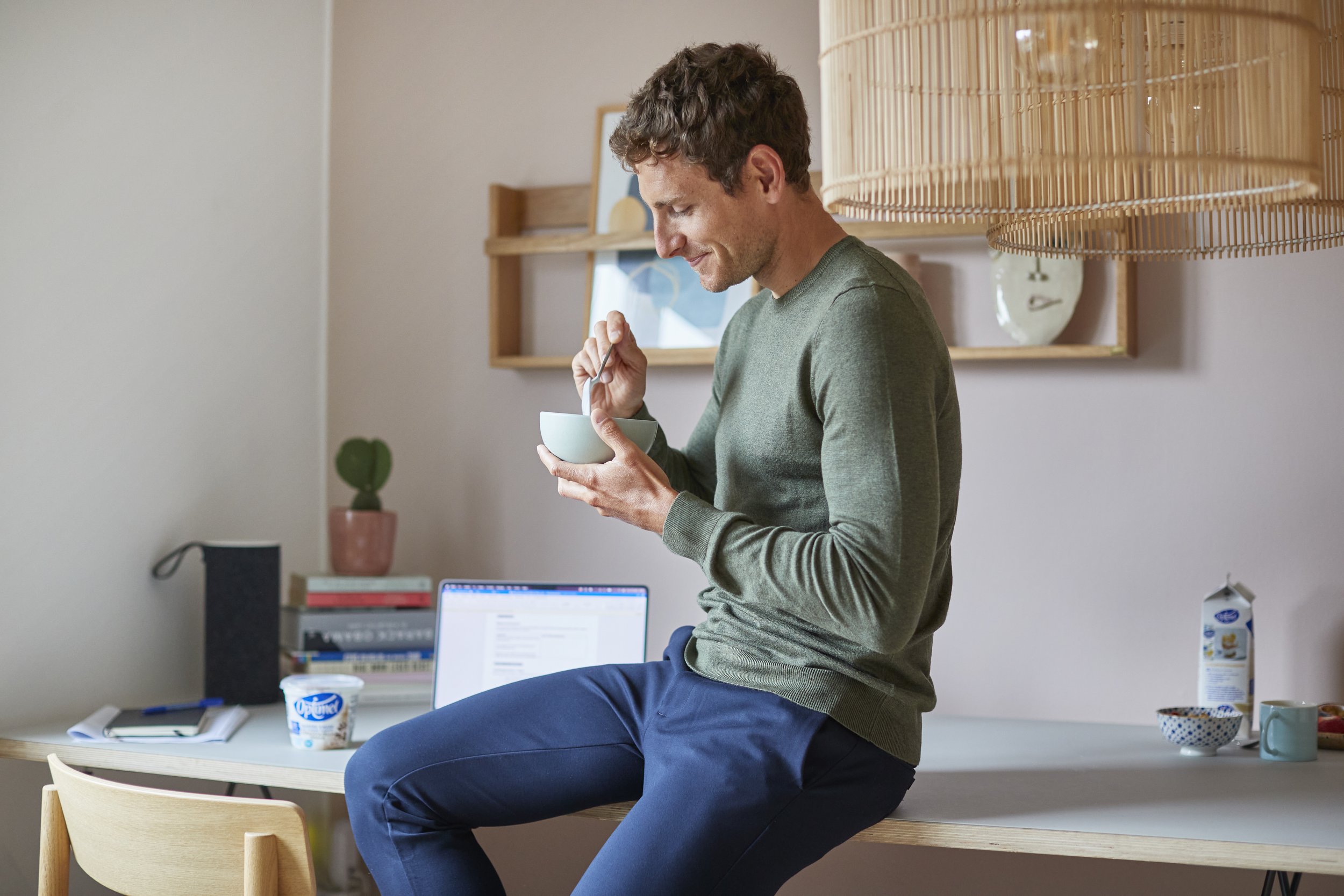 Mand sidder på en bordkant, drikker fra en skål med ske, i et hjemmekontor med computer, bøger, blomster, og morgenmad på bordet.