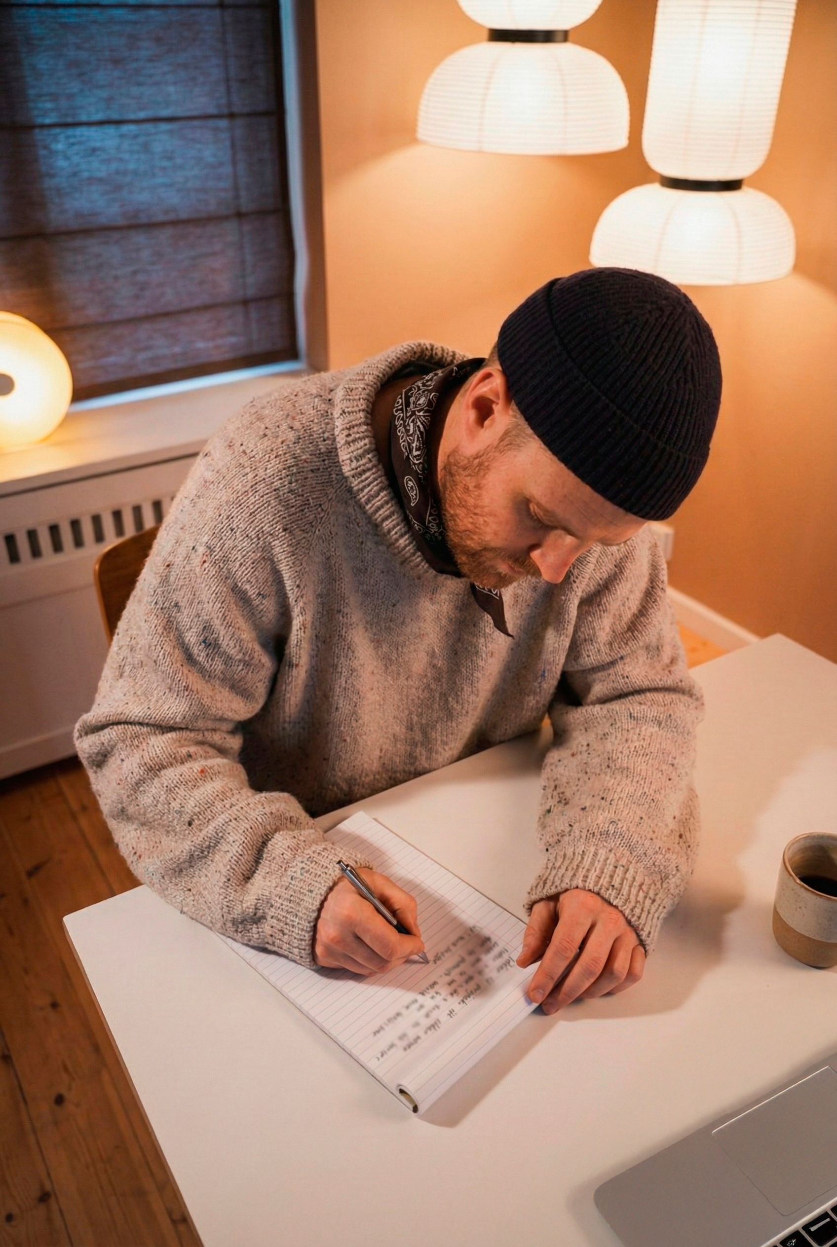 En mand skriver i en notesbog på et bord, med en kop kaffe ved siden af. Han har en sort hue og en grå sweater på, og der er bløde lamper i baggrunden.