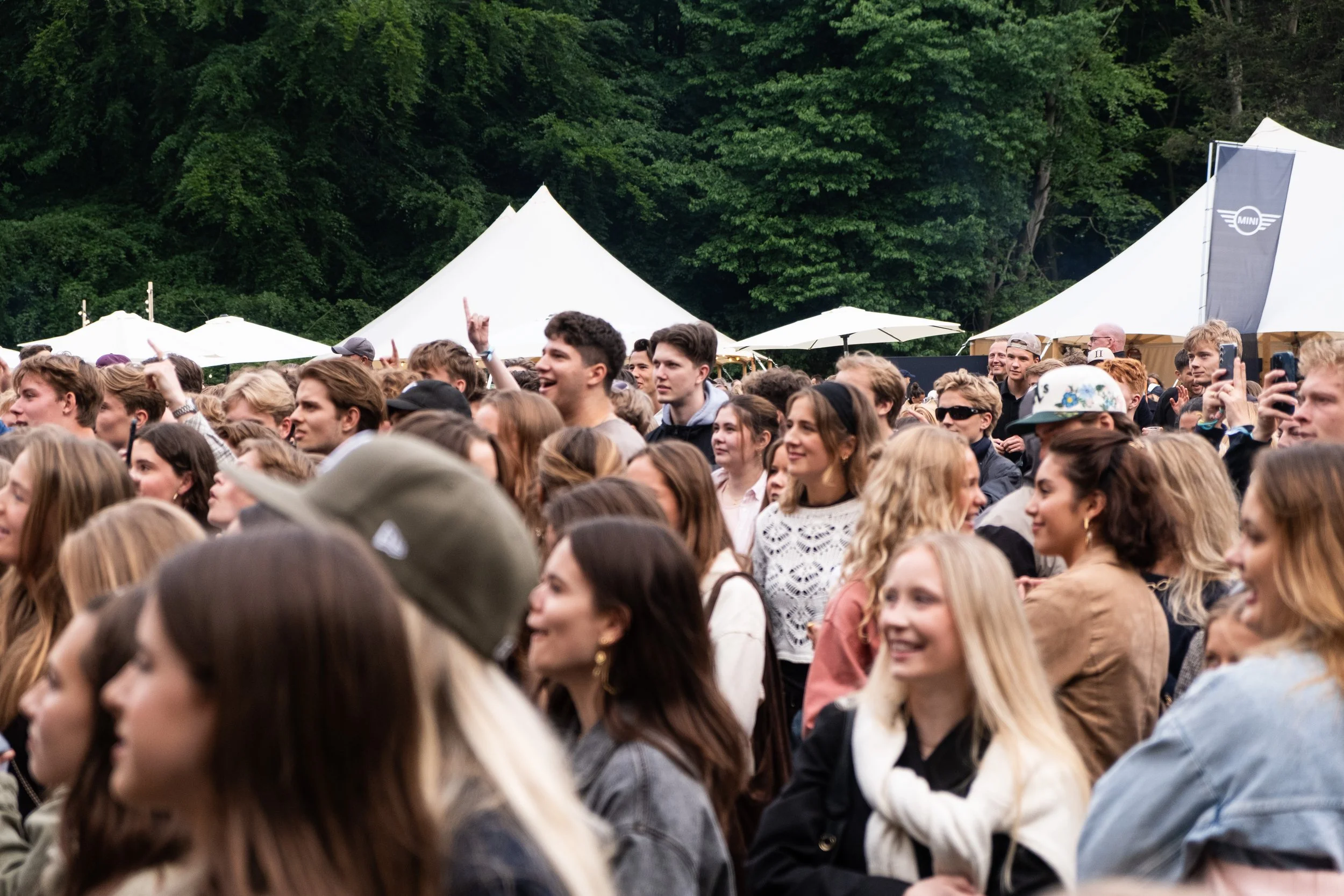 En stor folkemængde af unge mennesker, der nyder en koncert eller festival udendørs, med grønne træer i baggrunden og store hvide telte.