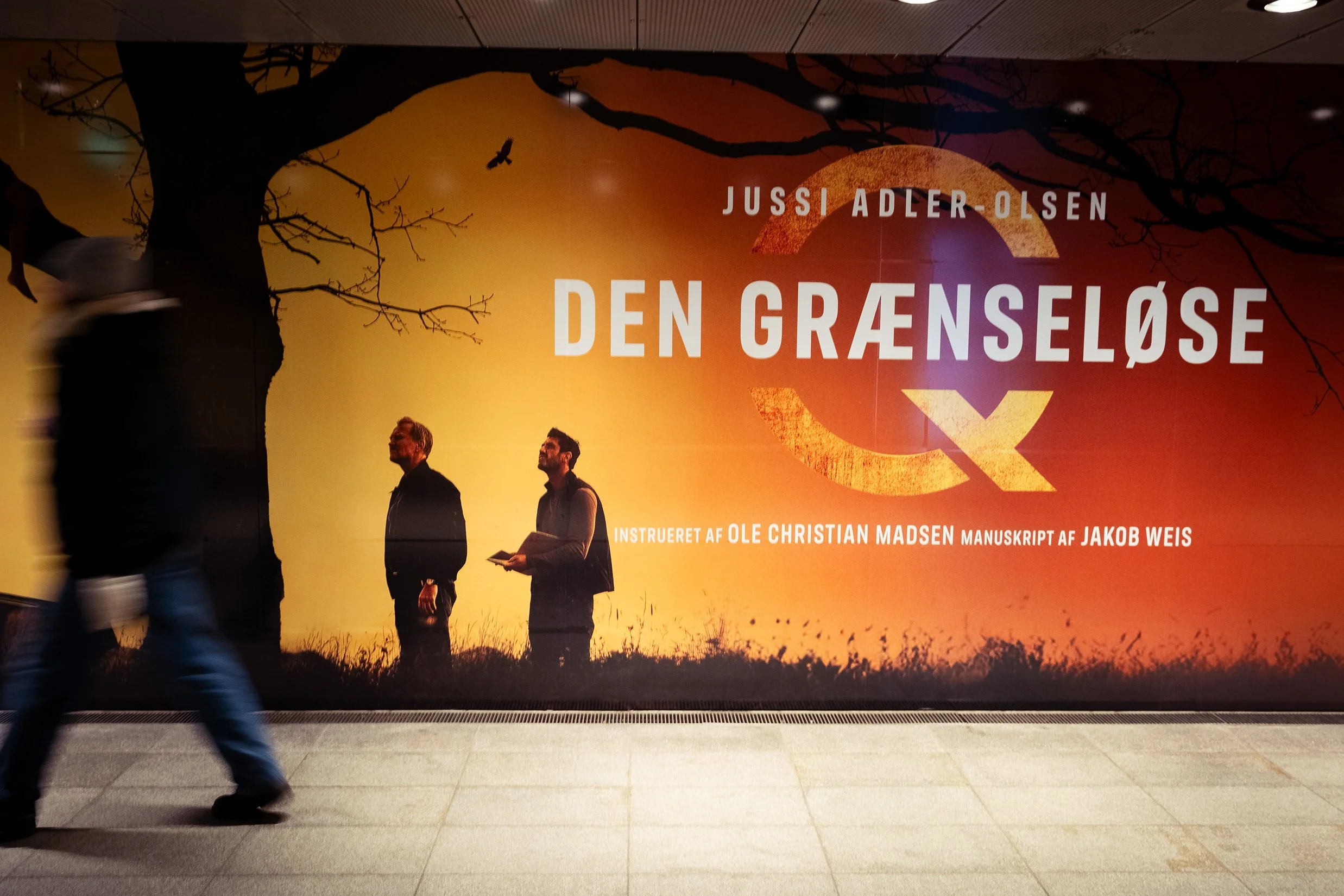 En reklamevægbeklædning for en teaterforestilling med titlen 'Den Grænseløse' med forfatteren Jussi Adler Olsen og instruktion af Ole Christian Madsen. Der ses tre personer i silhouet mod en baggrund af solnedgang eller solopgang, med en stor trægren