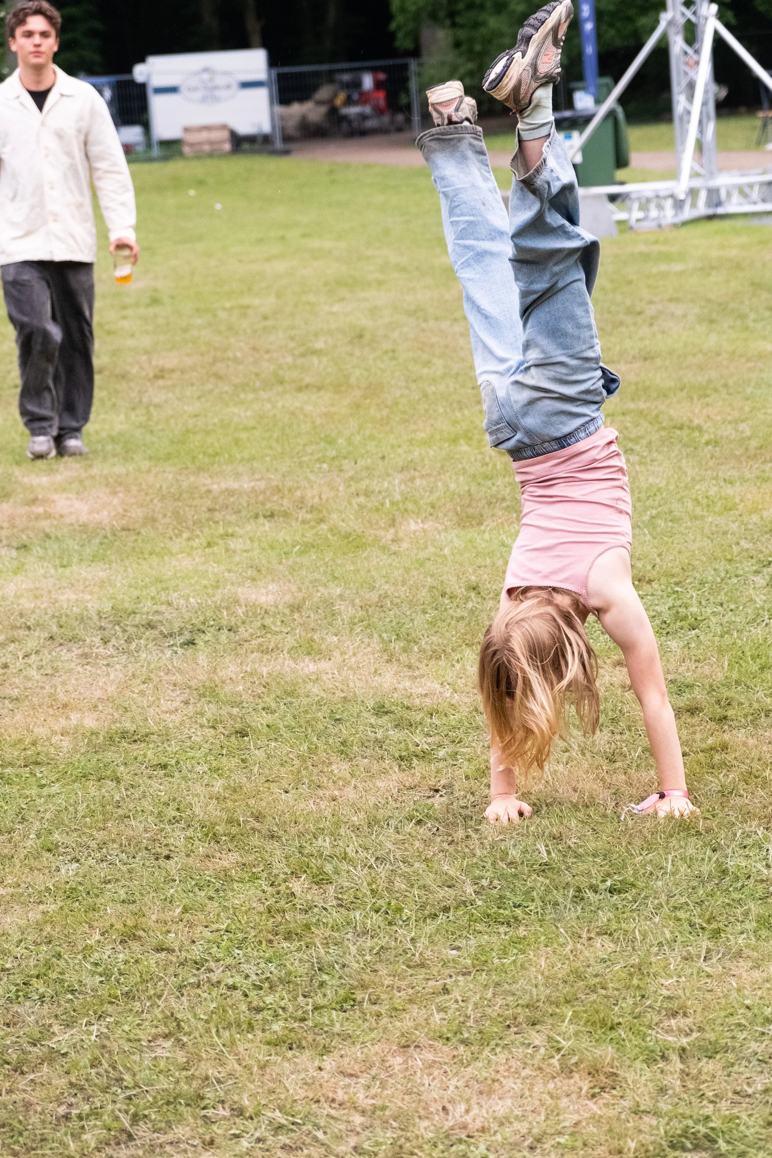 En pige laver handstand på græsplæne, mens en fyr holder en øl i baggrunden. Der er festlig udendørs stemning.