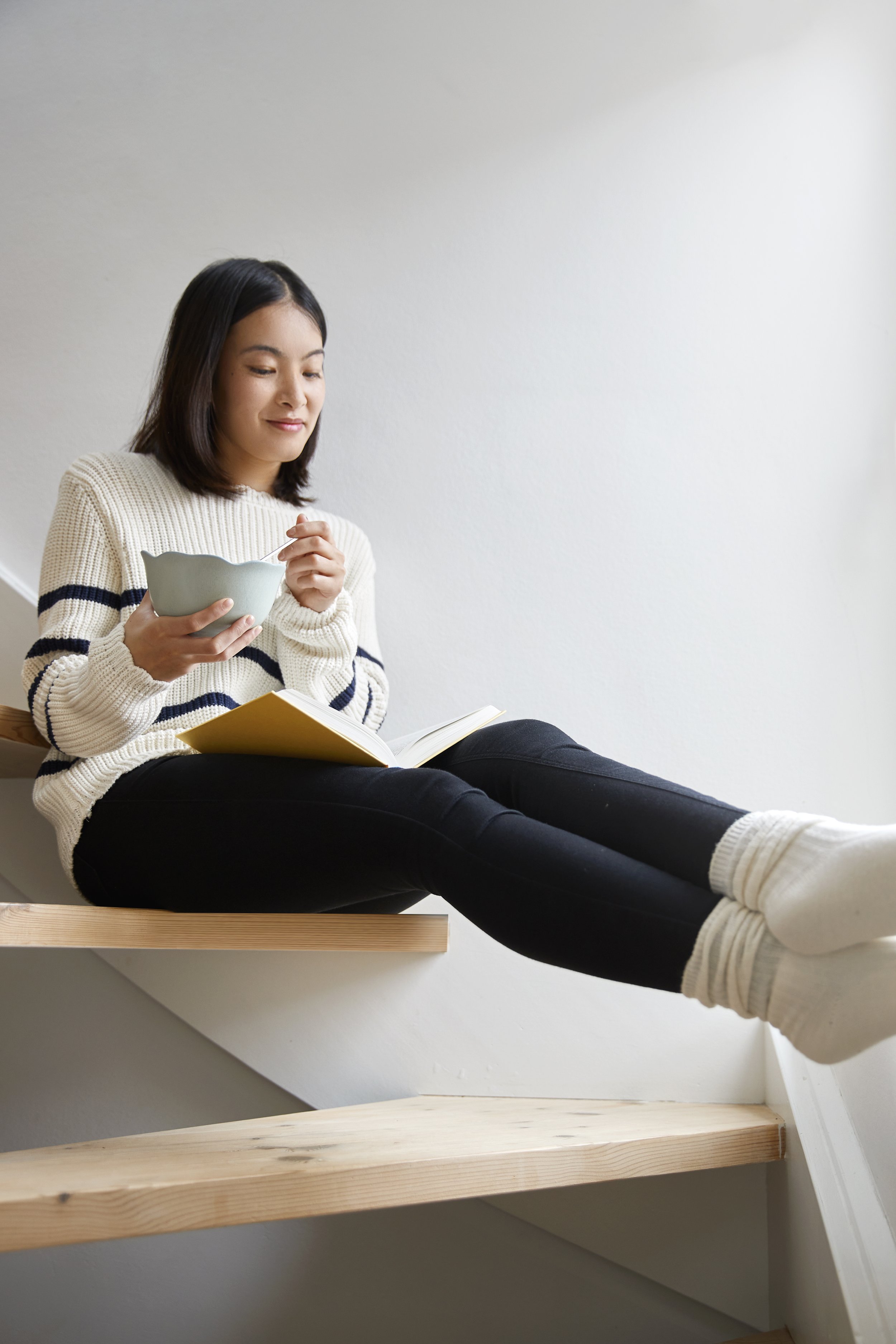 En ung kvinde sidder afslappet på en trappe, læser i en bog og spiser fra en skål.