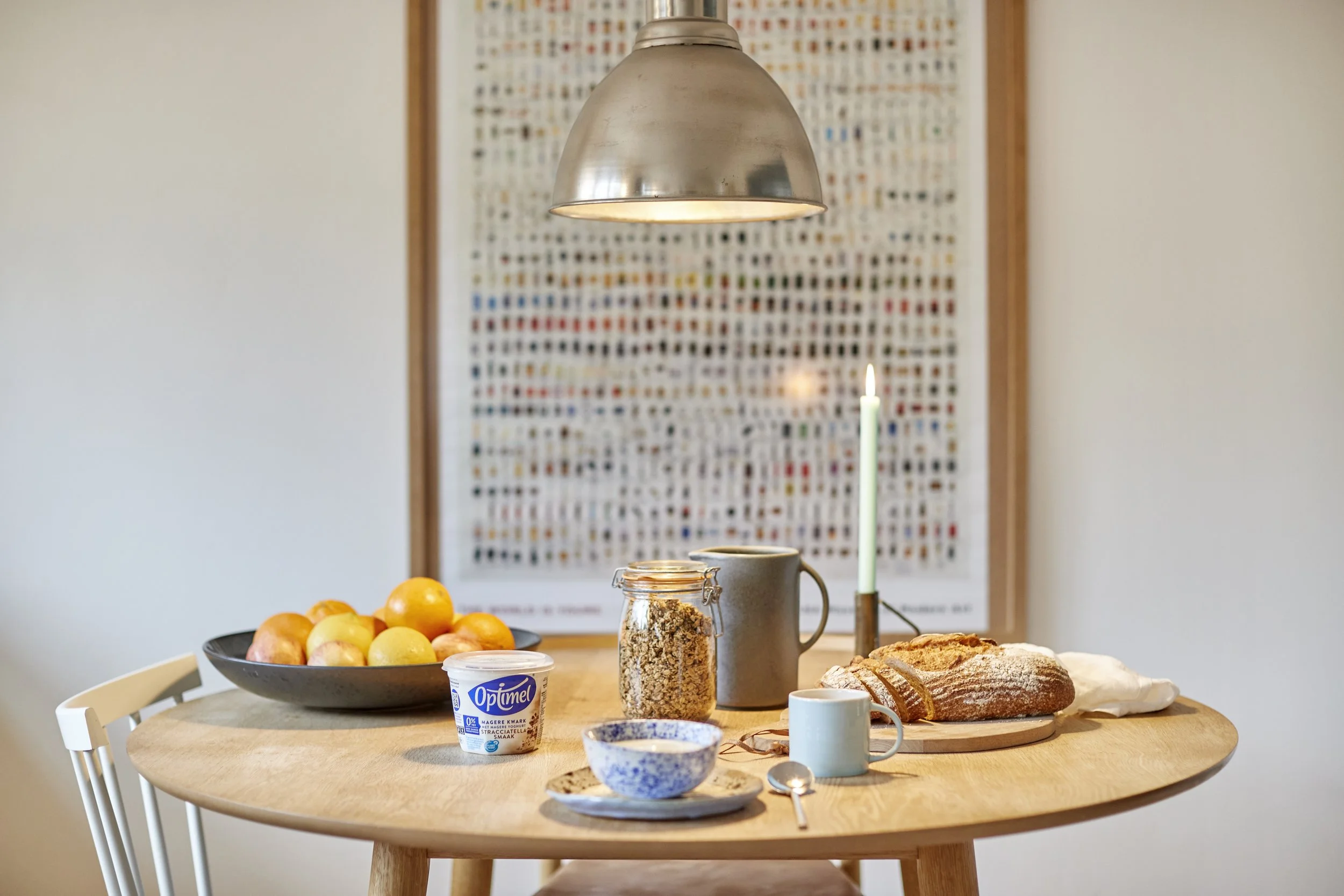 Madtab med frugt, yoghurt, granola, brød, ost, kopper, ske, lys, på et rundt træbord i et køkken.