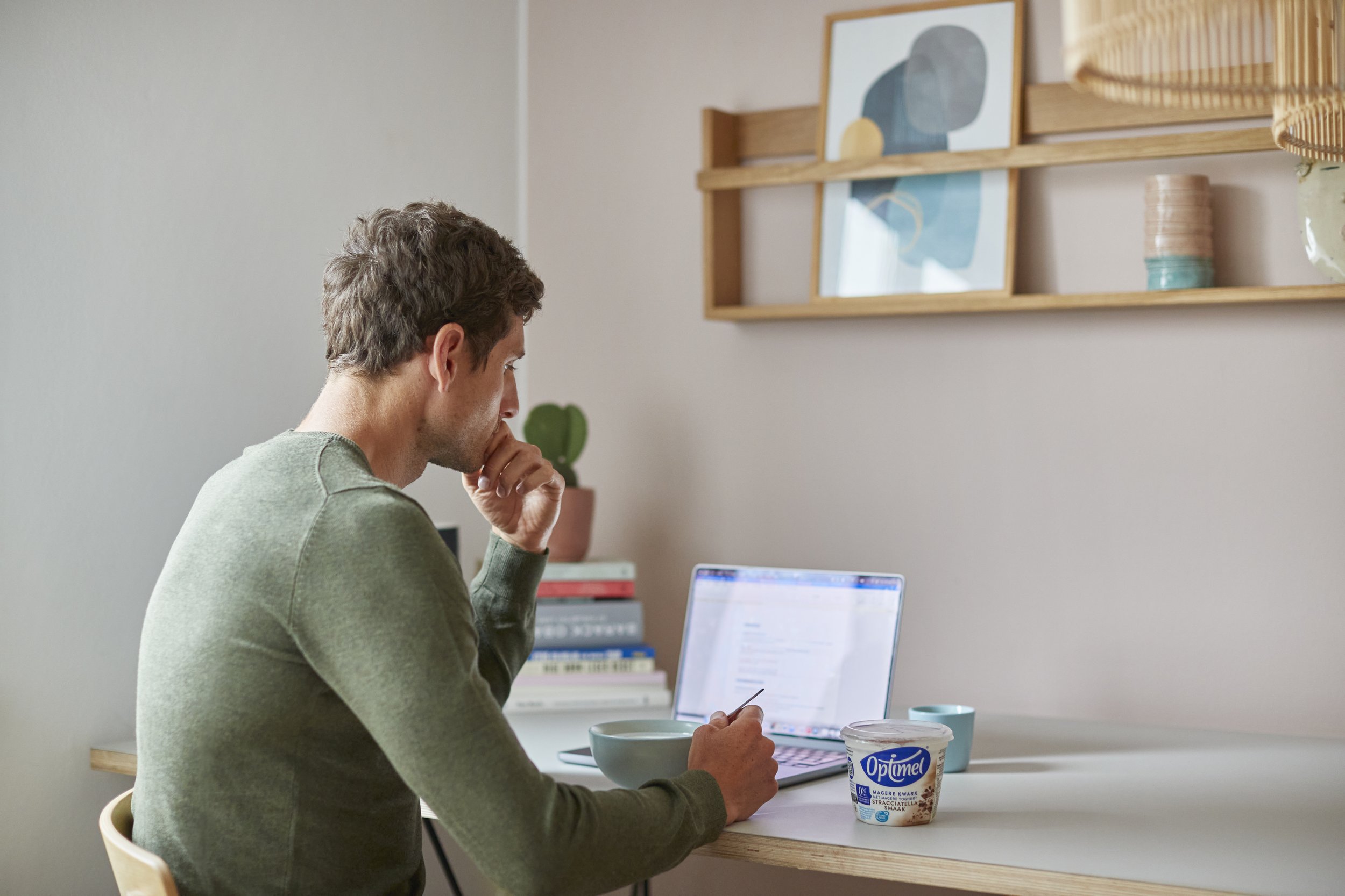 Mand sidder ved en hvid skrivebord med en åben laptop, en skål, en bæger is, og en kop, foran ham er der også en books og en plante, mens han arbejder eller læser, i en enkel, moderne værelse.