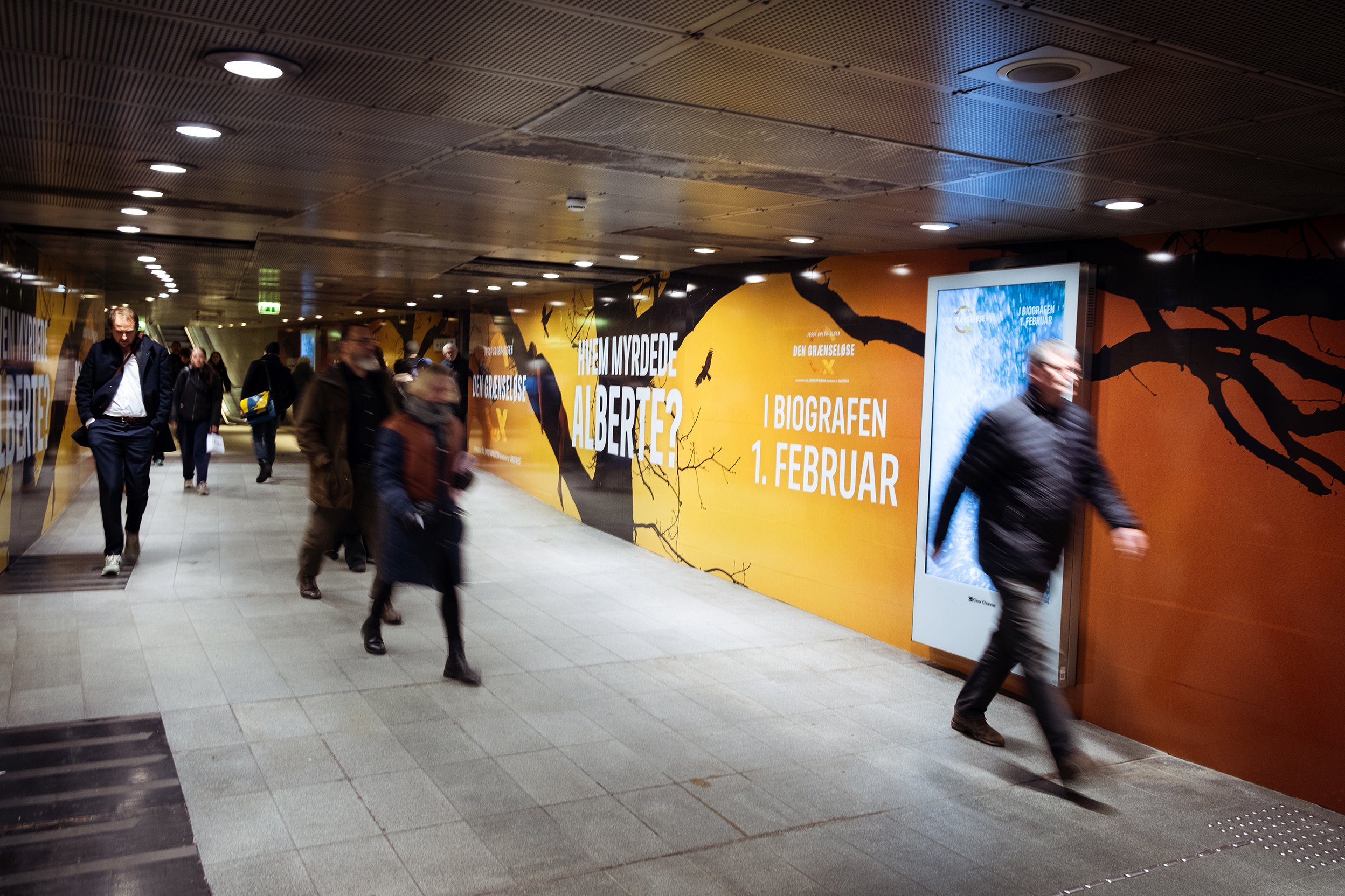 Folk går gennem en underjordisk gang med reklamer på væggene, der annoncerer en biograffilm premiere den 1. februar.