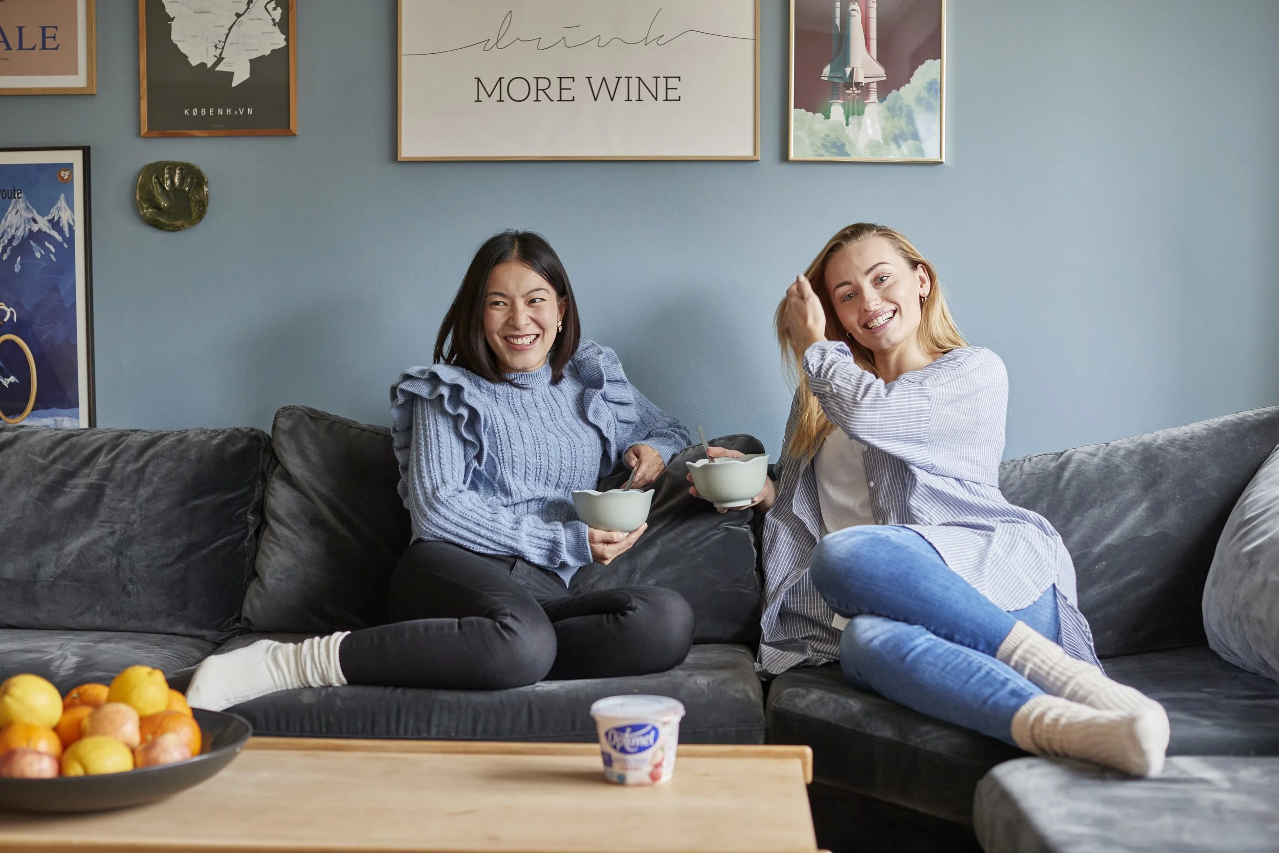 To kvinder sidder på en sofa og nyder yoghurt, mens de smiler til kameraet. Der er en skål med æbler på et bord foran dem.