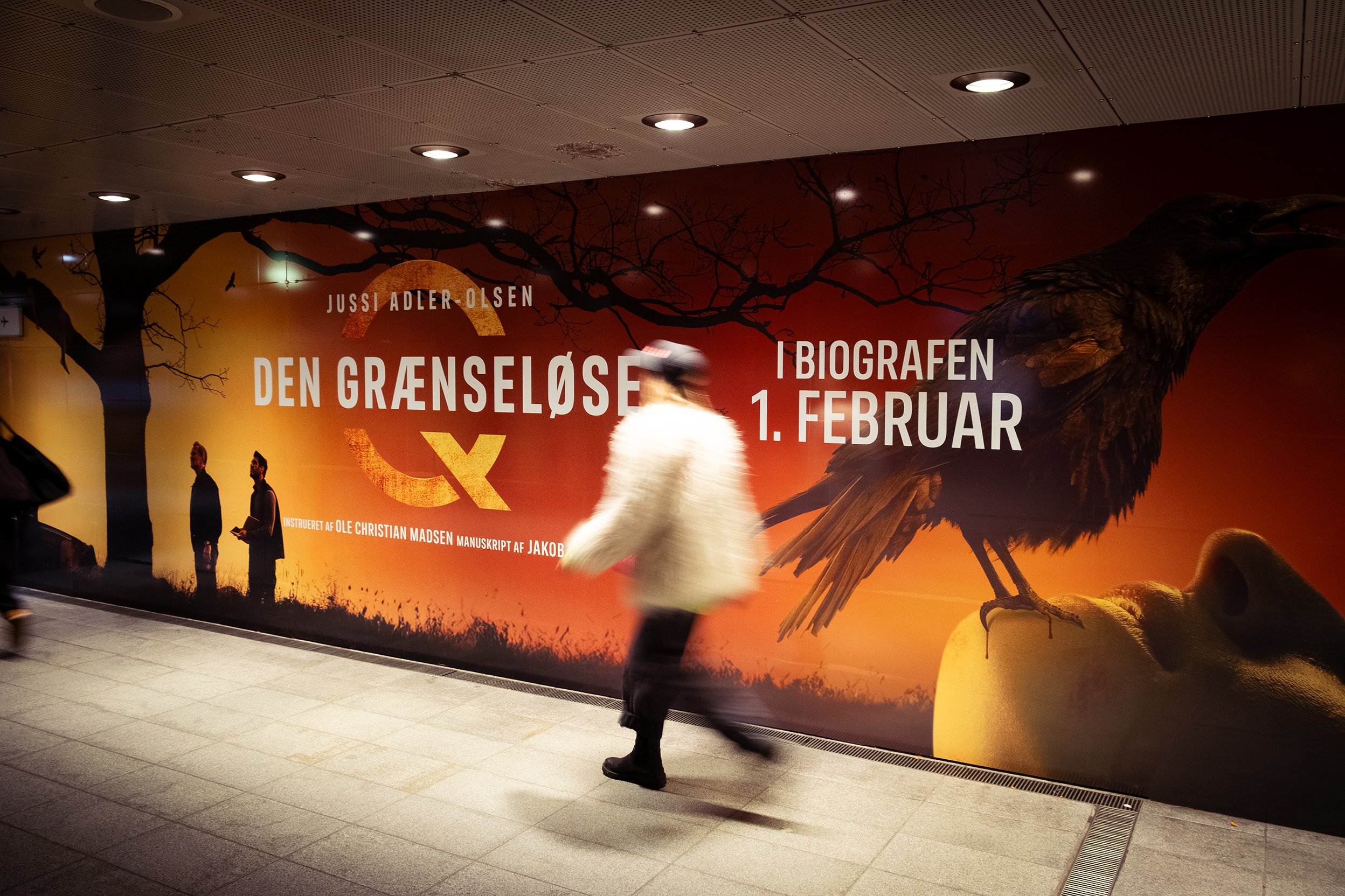 En person går gennem en undergrundsstation med en stor reklameplakat på væggen for en film kaldet 'Den Grænseløse', der åbner i biografen den 1. februar. Reklameplakaten har en fugl og spilende træer i baggrunden.