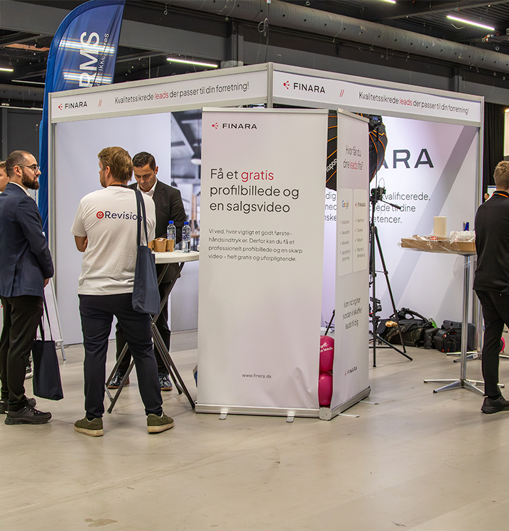 En messeudstilling med reklameskilte for Finara og Revisionsfirma, hvor personer snakker og står omkring standen.