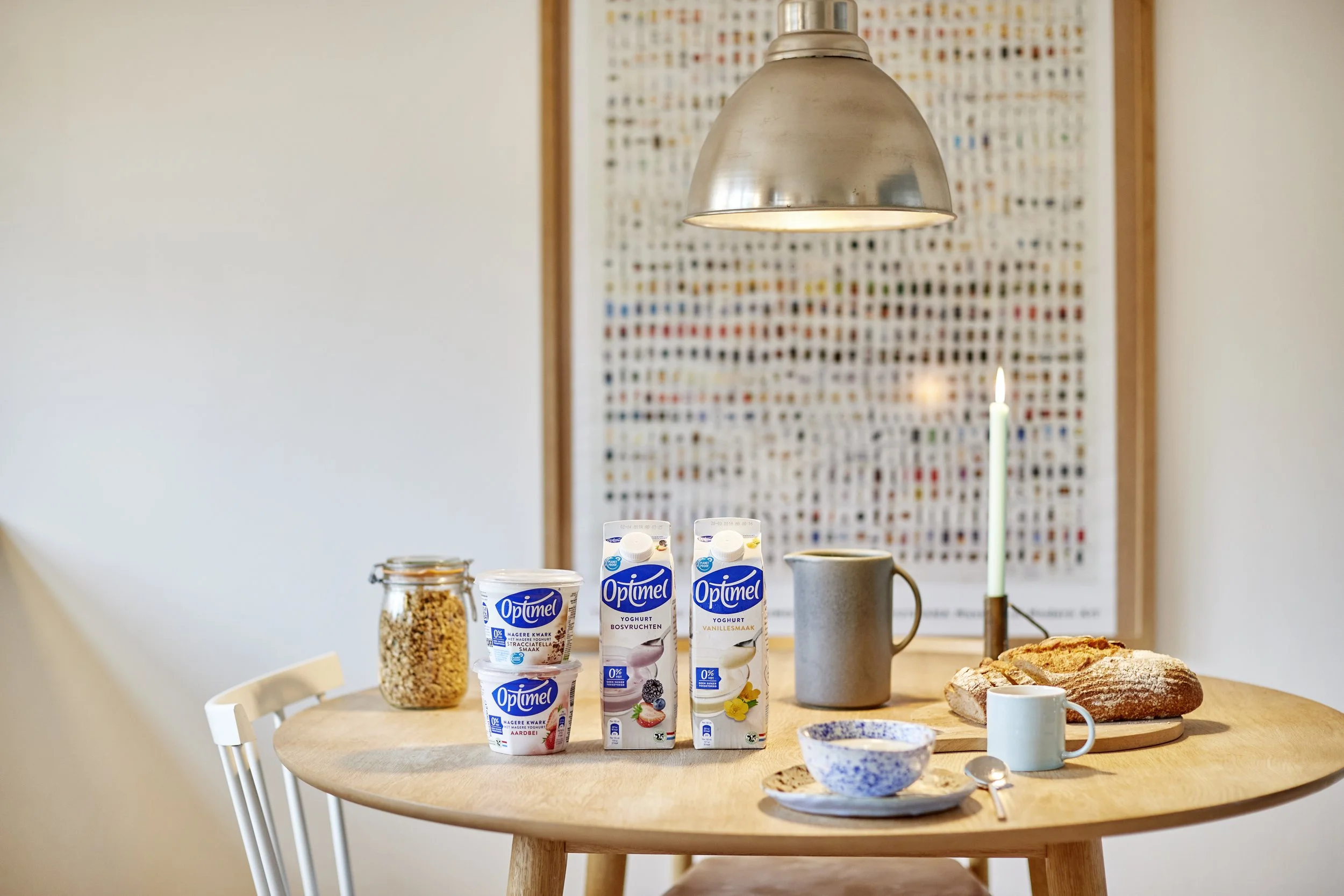 Et rundt træbord med yoghurt, brød, kopper, ske, julelys, og en baggrund med et farverigt vægbillede.