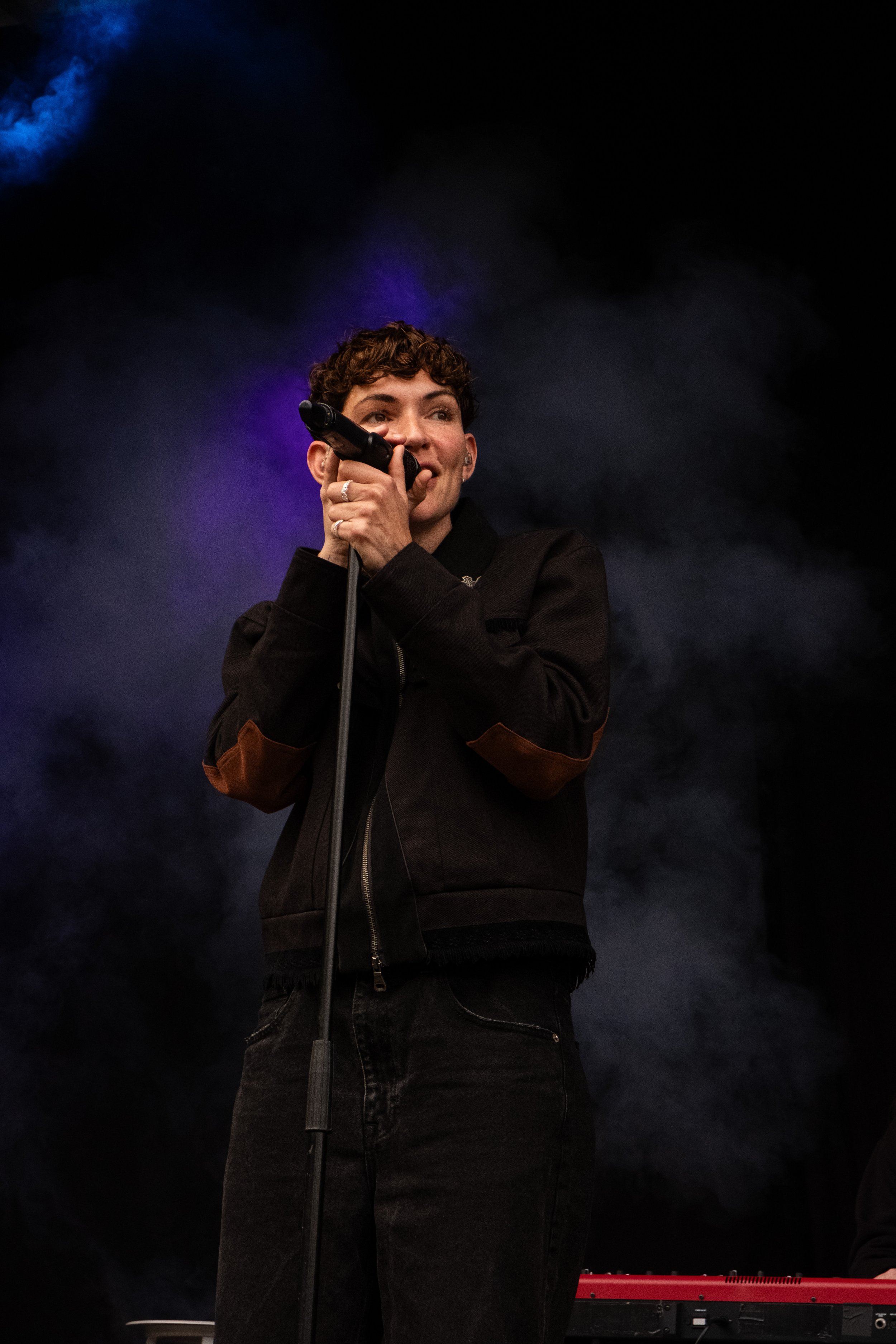 Kvinde med kort, krøllet hår synger ind i en mikrofon på scenen med mørk baggrund og lidt røg.