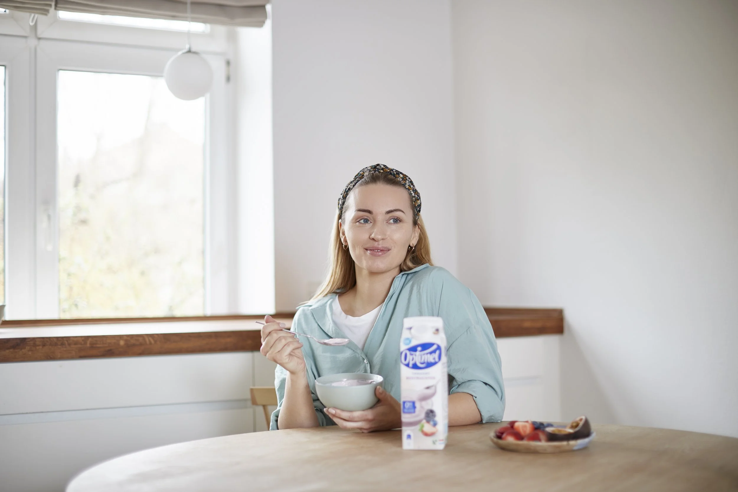 Kvinde sidder ved et bord med en bowl yoghurt, en karton yoghurt og en tallerken med frugt, spiser yoghurt og smiler.