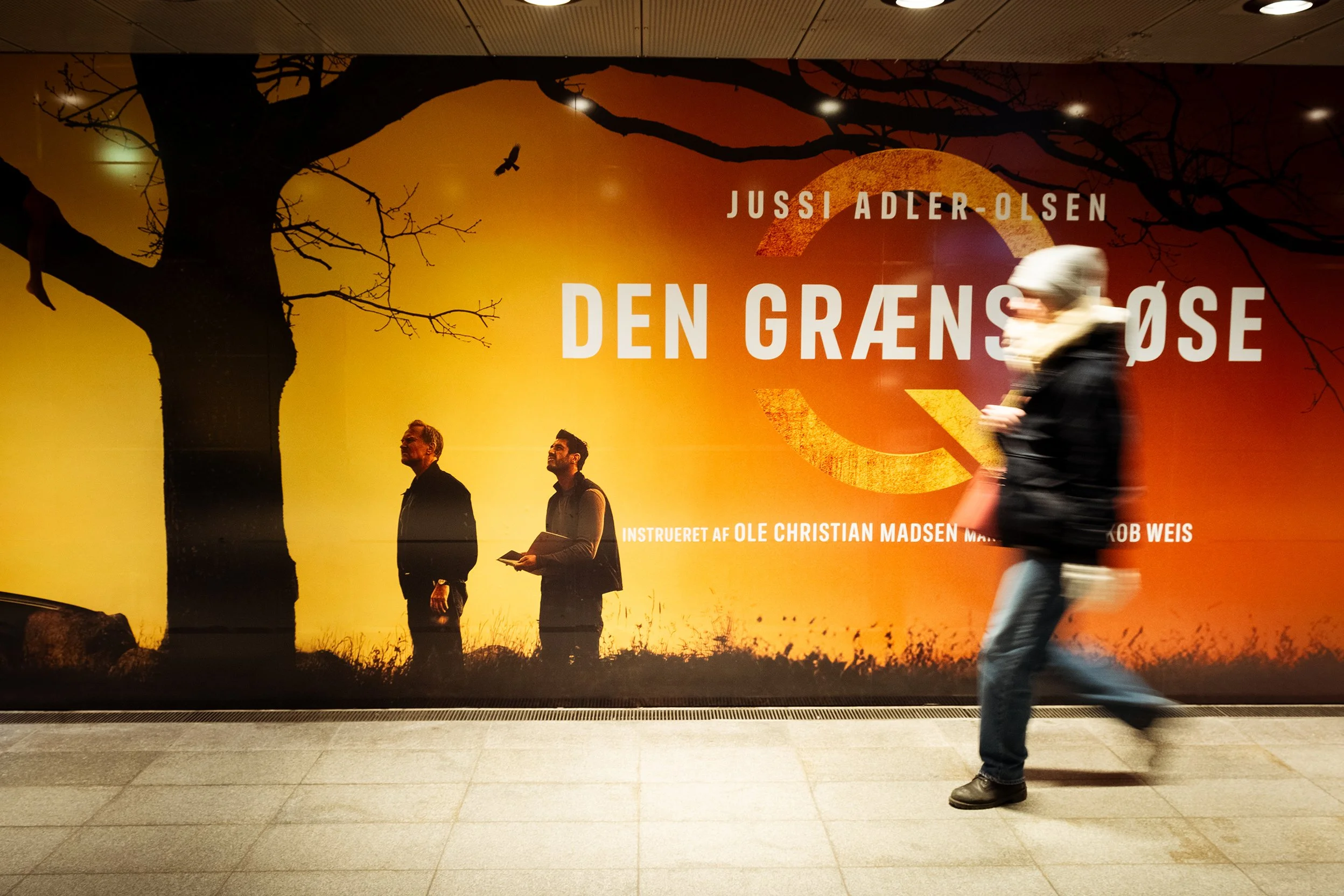 En person går forbi en reklamevæg foran en metrostation, reklamen er for en teaterforestilling med titlen 'Den Grænseløse' af Jussi Adler-Olsen. Væggen har en stor, sort silhuet af et træ med nogle grene uden blade, og en sol, der er delvist dækket a